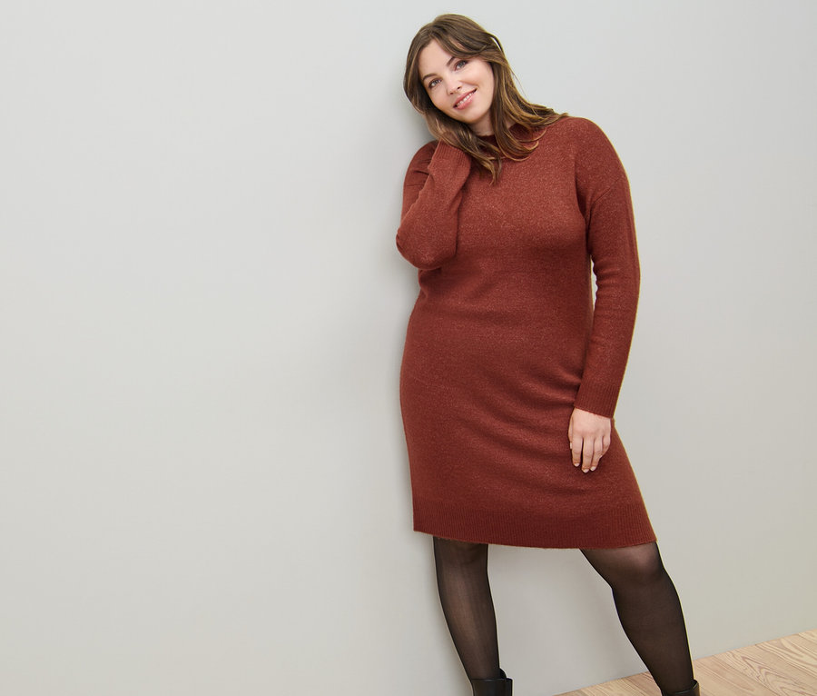 Egy nő a falnak dőlve kötött ruhát visel gyapjúval és egy fekete, 50 DEN-es, átlátszó hatású harisnyát.