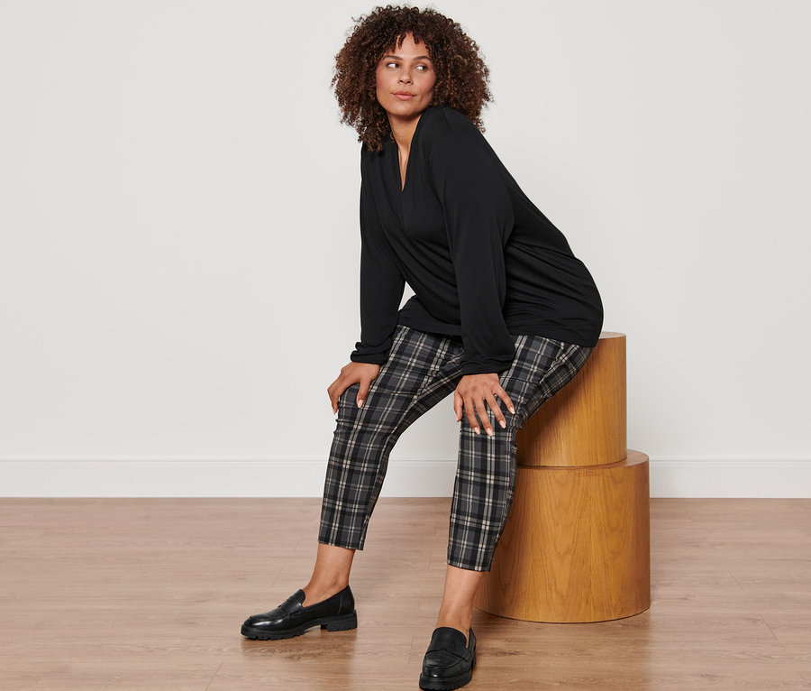 Egy nő ül egy fa ülőkén, fekete V-nyakú hosszú ujjú felsőt, kockás sztreccsnadrágot és bőr mokaszint visel.