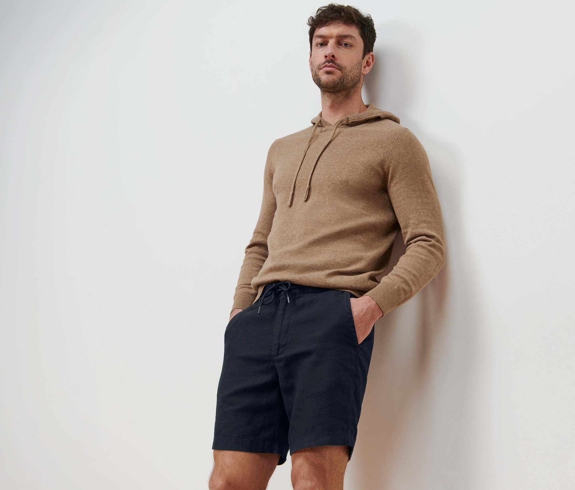 Férfi a falnak dőlve, lenvászon rövidnadrágot és kapucnis pulóvert visel.