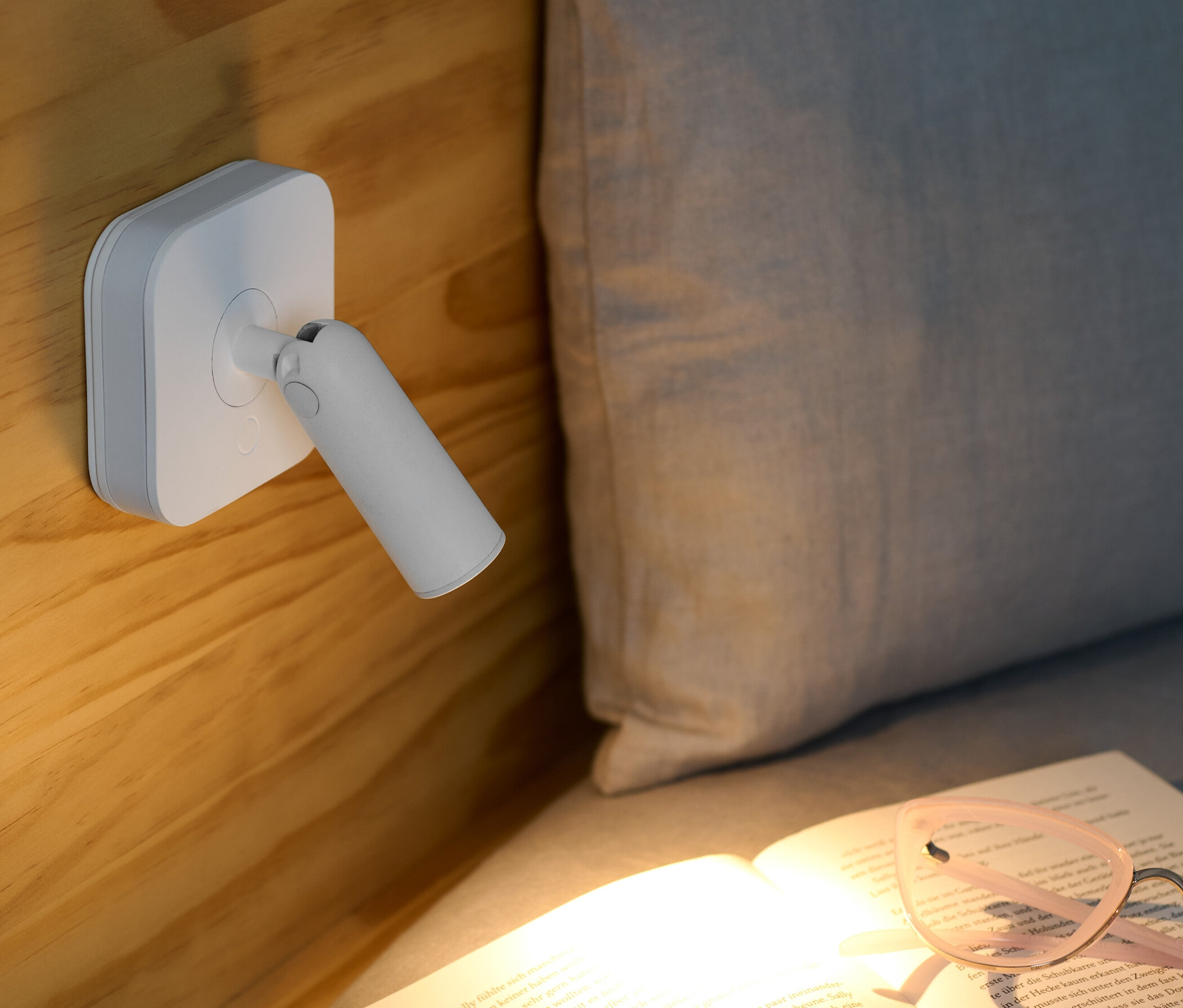 A fából készült ágyhoz egy LED-es éjjeli lámpa van rögzítve, spotlámpával, amely egy nyitott könyvet világít meg, rajta egy szemüveggel.