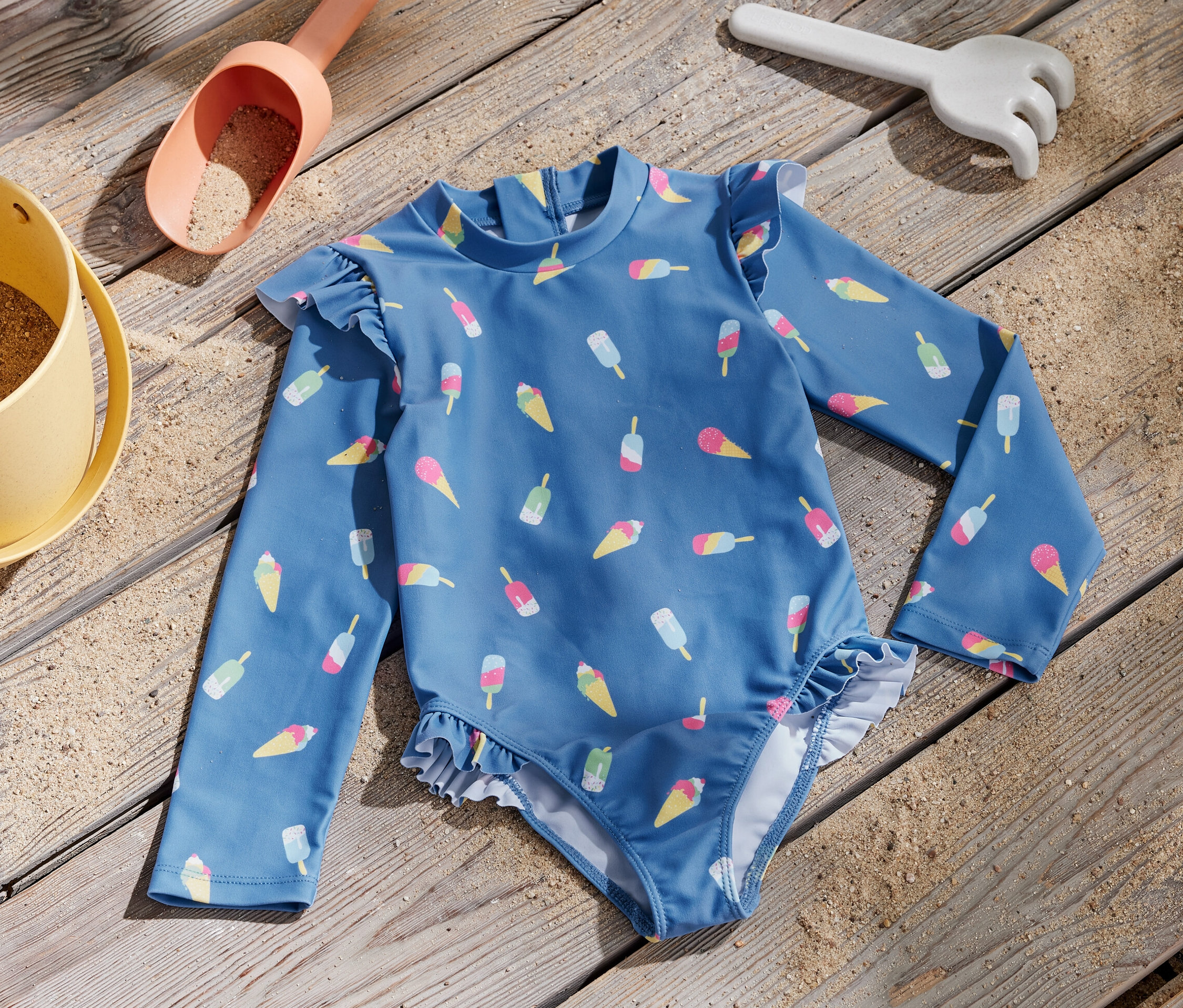 Kék, fagylalt mintás gyerek fürdőruha 80-as UV védelemmel, egy fa deszkán homokozó játékok mellett.