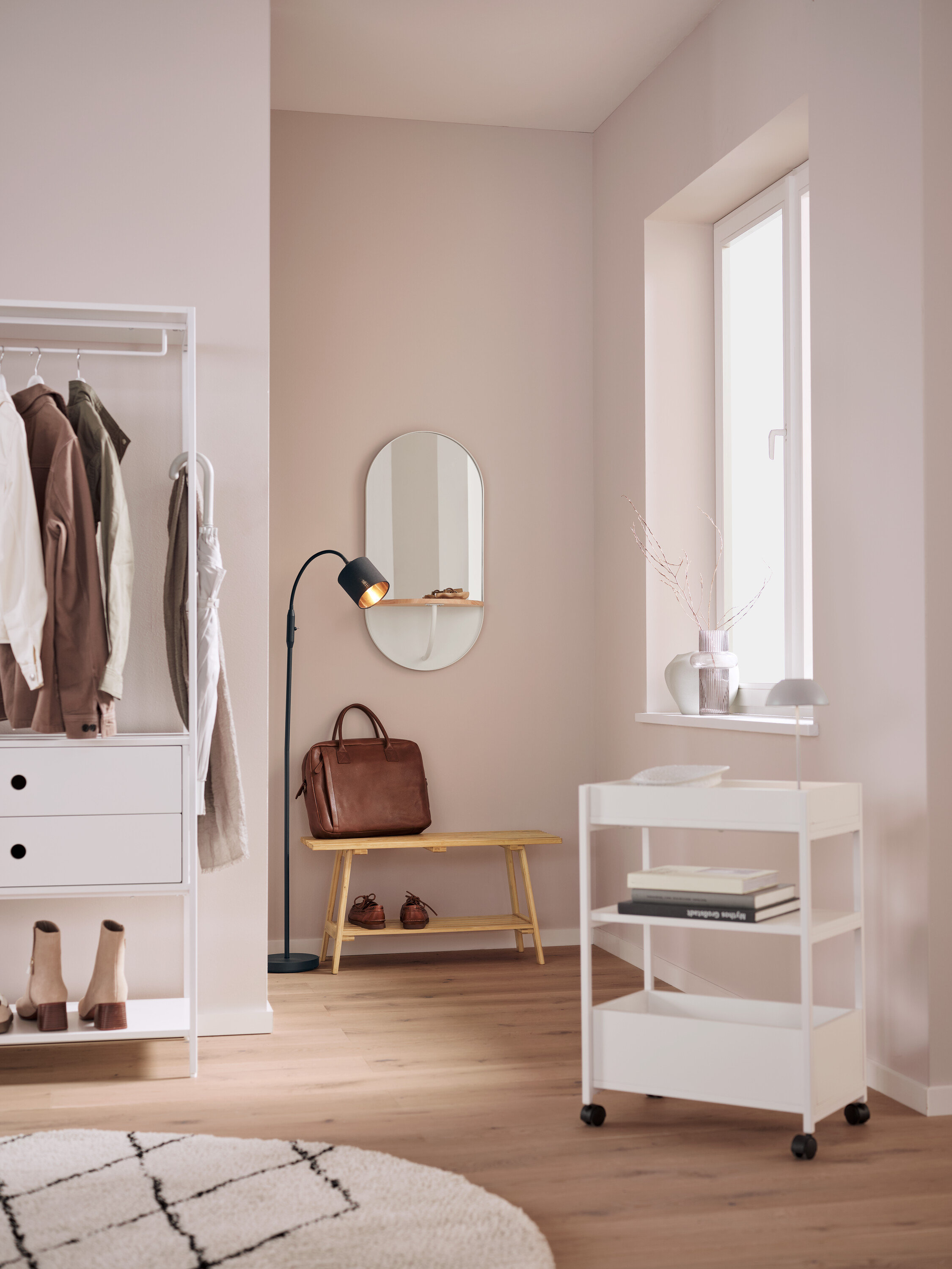 Egy belső térben egy fehér polc ruhákkal, egy ovális tükör, egy barna bőrtáska egy padon, egy fekete lámpa és egy fehér gurulós kocsi található.