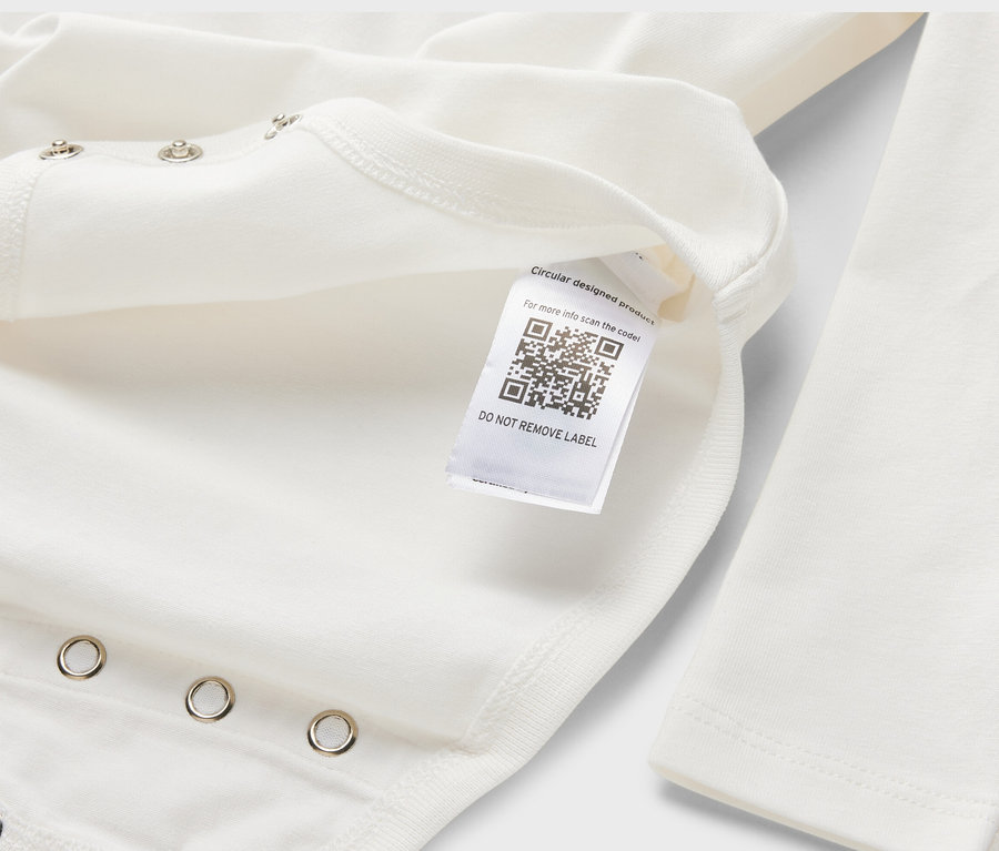 Egy fehér, hosszú ujjú, állítható hosszúságú kisgyerek body részlete, QR-kóddal ellátott címkével.