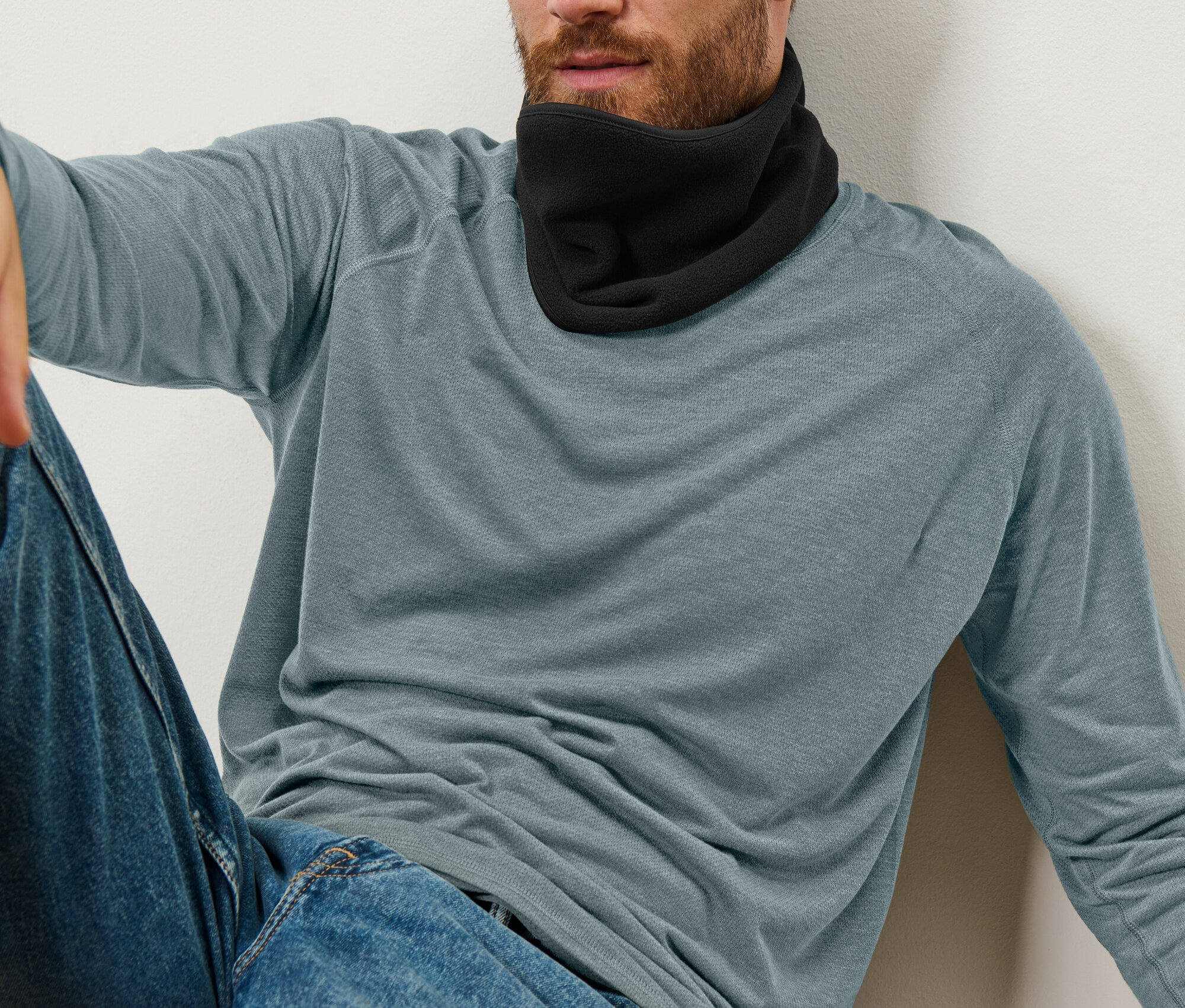 Egy férfi ül a falnak dőlve, fekete nyakmelegítőt, kék farmert és kék-szürke hosszú ujjú pólót visel.