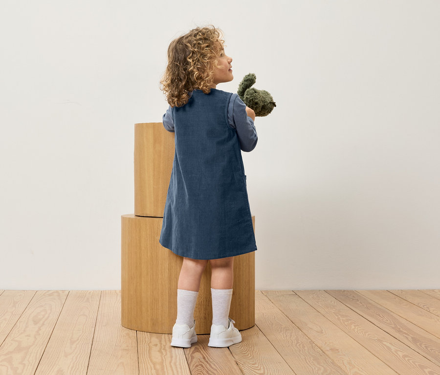 Egy kék ruhás lány plüssjátékot tart a kezében, és fa kockák mellett áll.