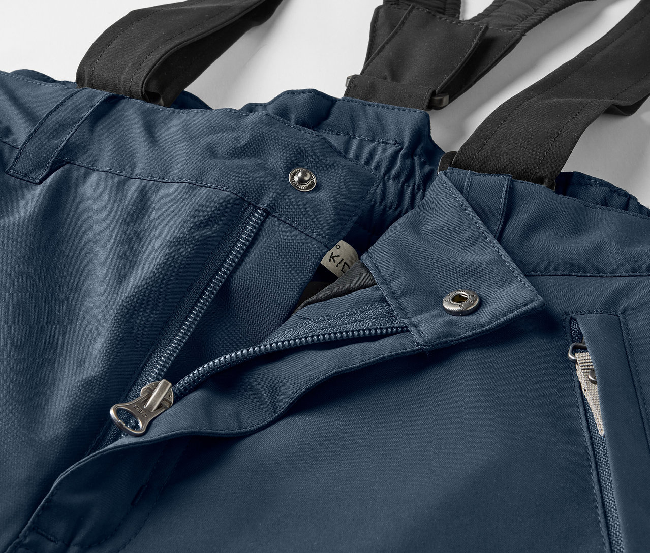 Kék nadrág részlete fekete nadrágtartókkal, nyitott cipzárral és ezüst gombokkal.