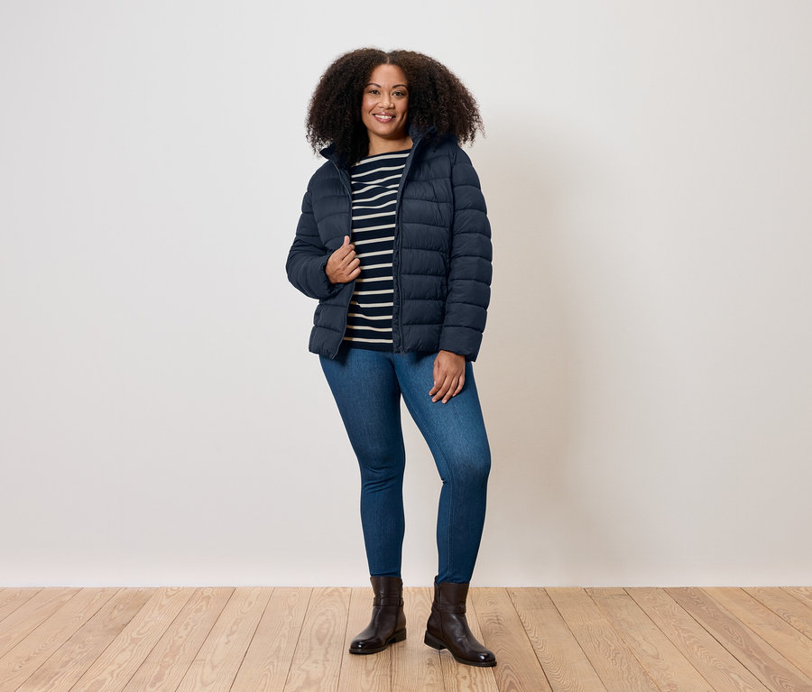 Egy göndör hajú nő áll, kék steppelt kabátot, csíkos pólót, farmert és csizmát visel.