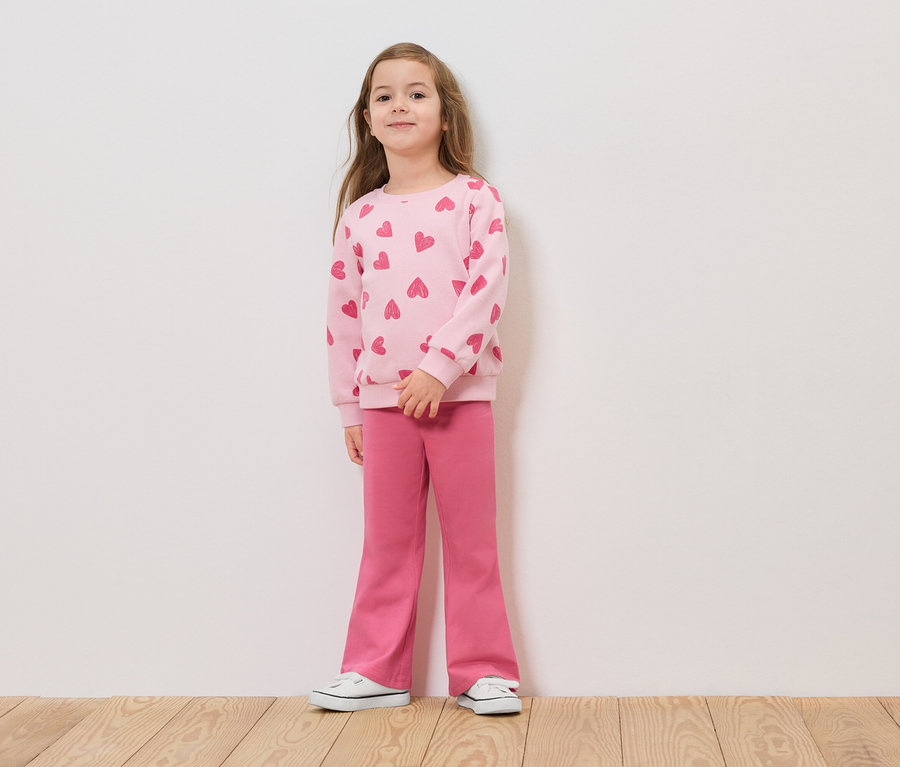 Egy lány áll egy fehér fal előtt, rózsaszín, szívecskés pulóverben és rózsaszín nadrágban.