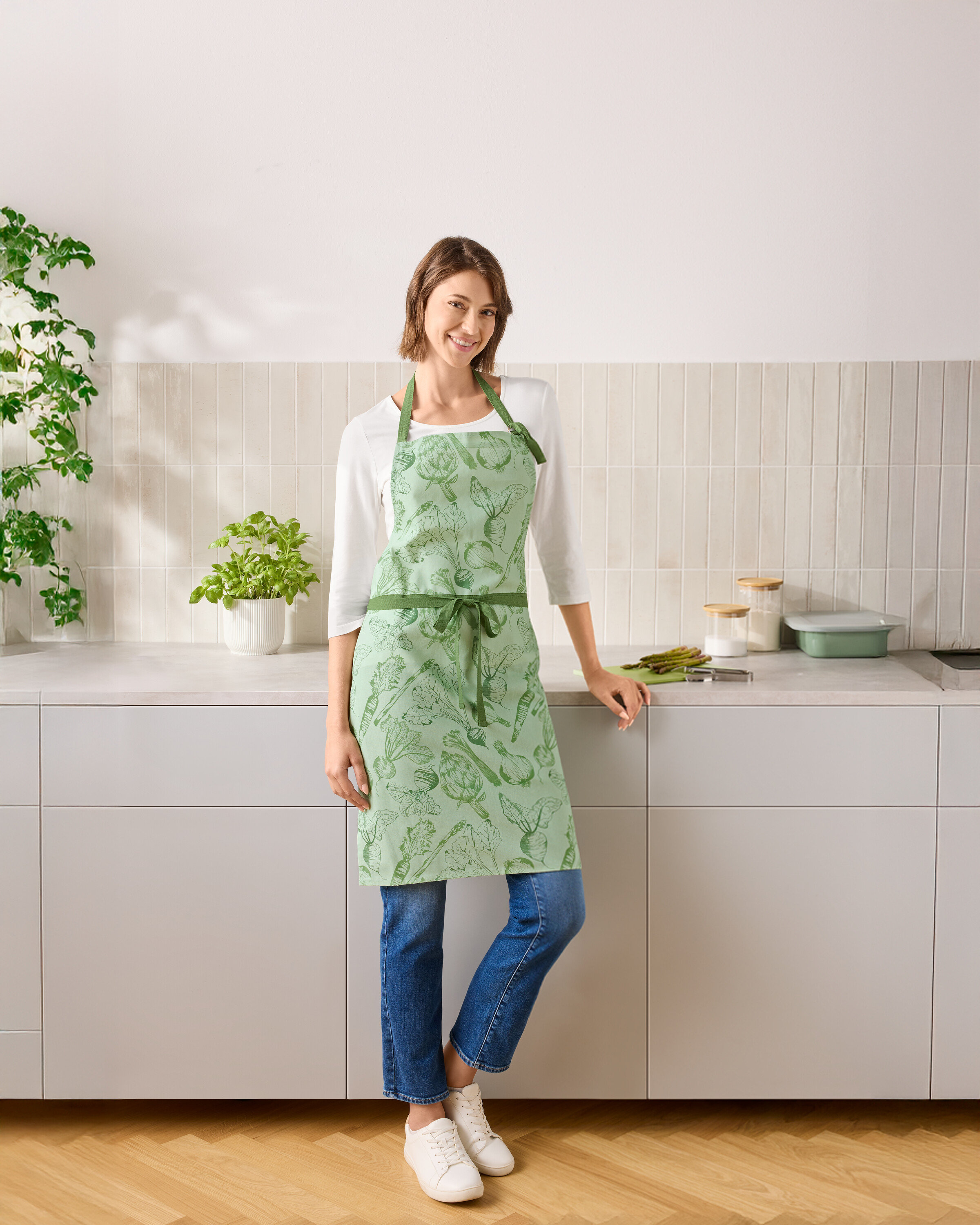 Egy nő áll a konyhában, zöld, zöldséges kötényt visel.