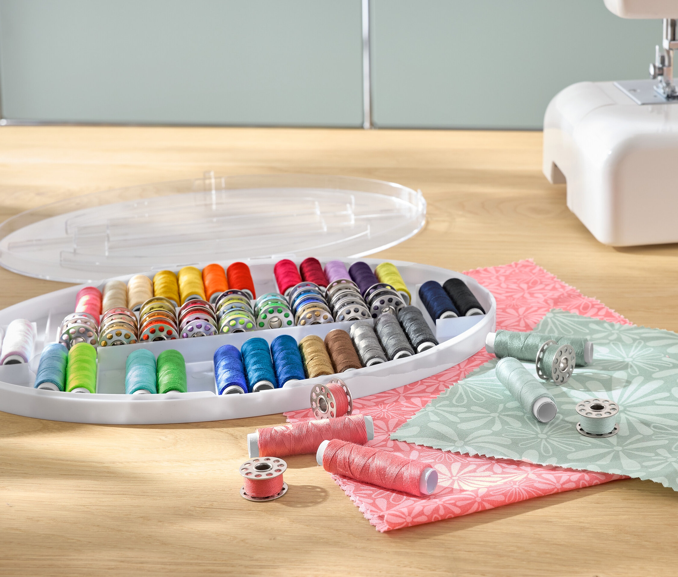 Egy színes cérnakészlet orsókon egy műanyag tárolóban, egy varrógép és szövetek mellett.
