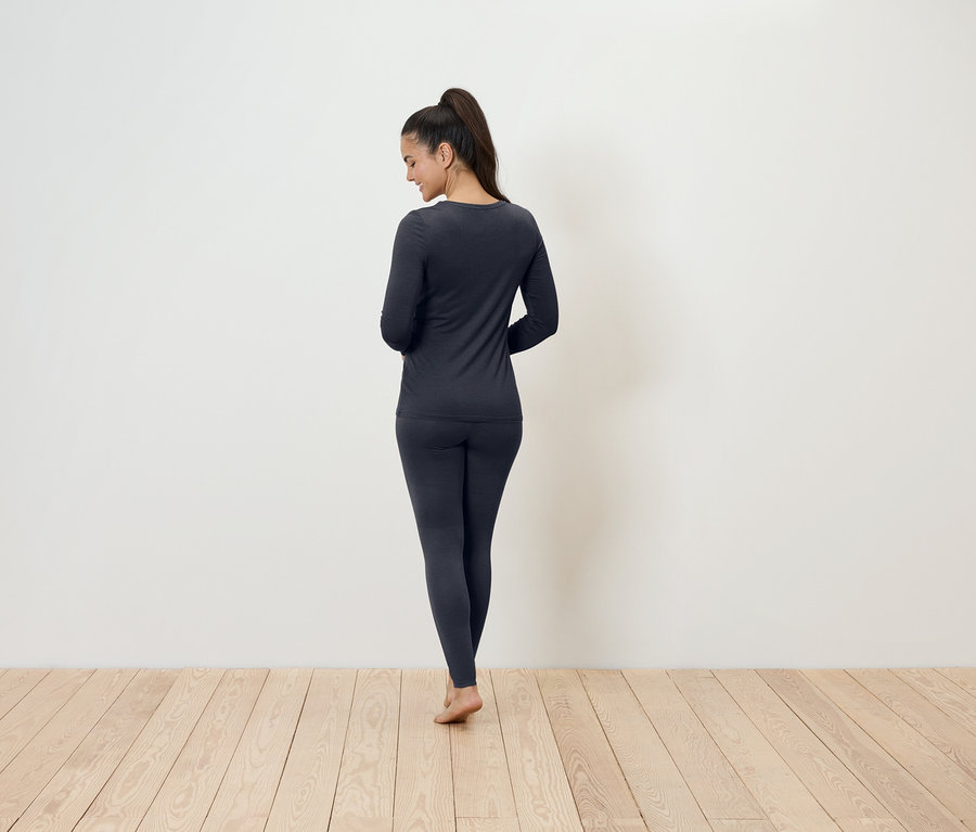 Egy nő áll háttal a kamerának, sötétszürke termikus fehérneműt visel.