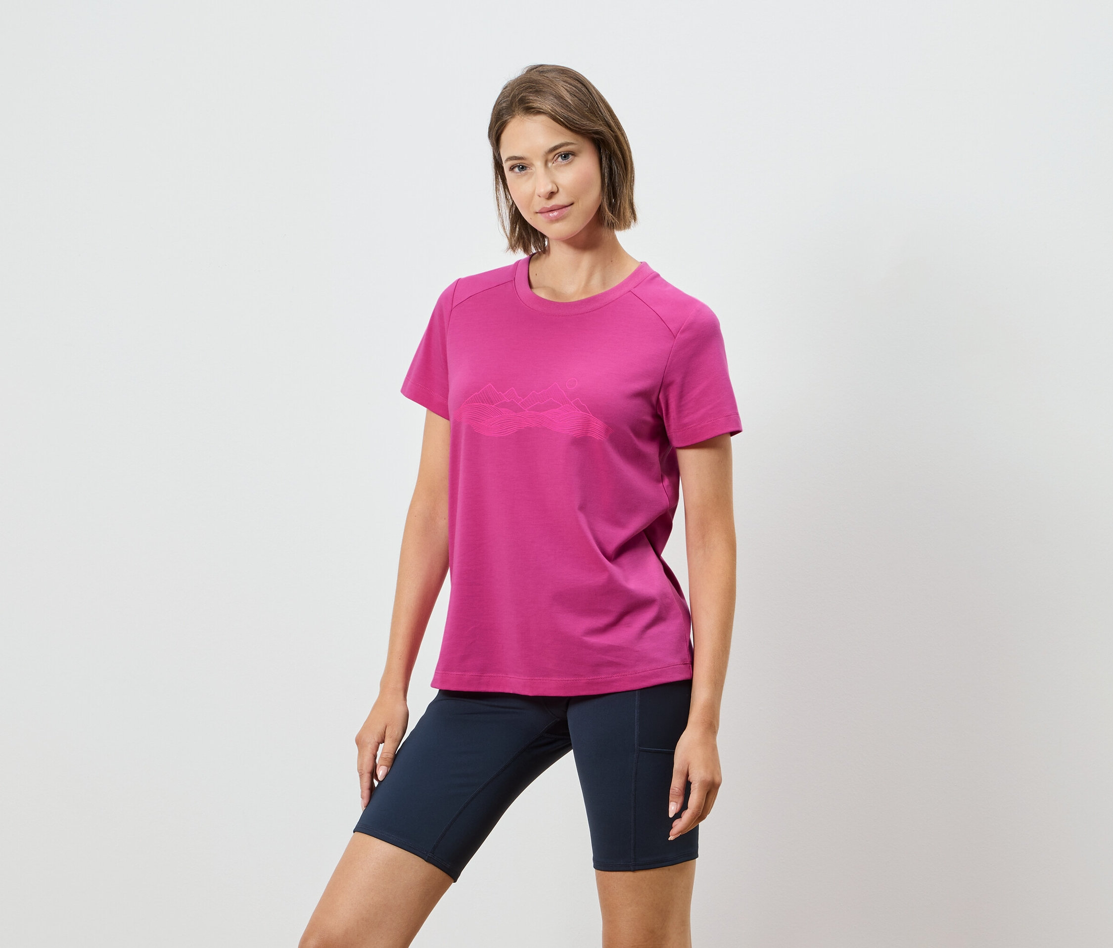 Egy nő rózsaszín pólót visel hegyvonulattal és sötétkék rövidnadrágot fehér háttér előtt.