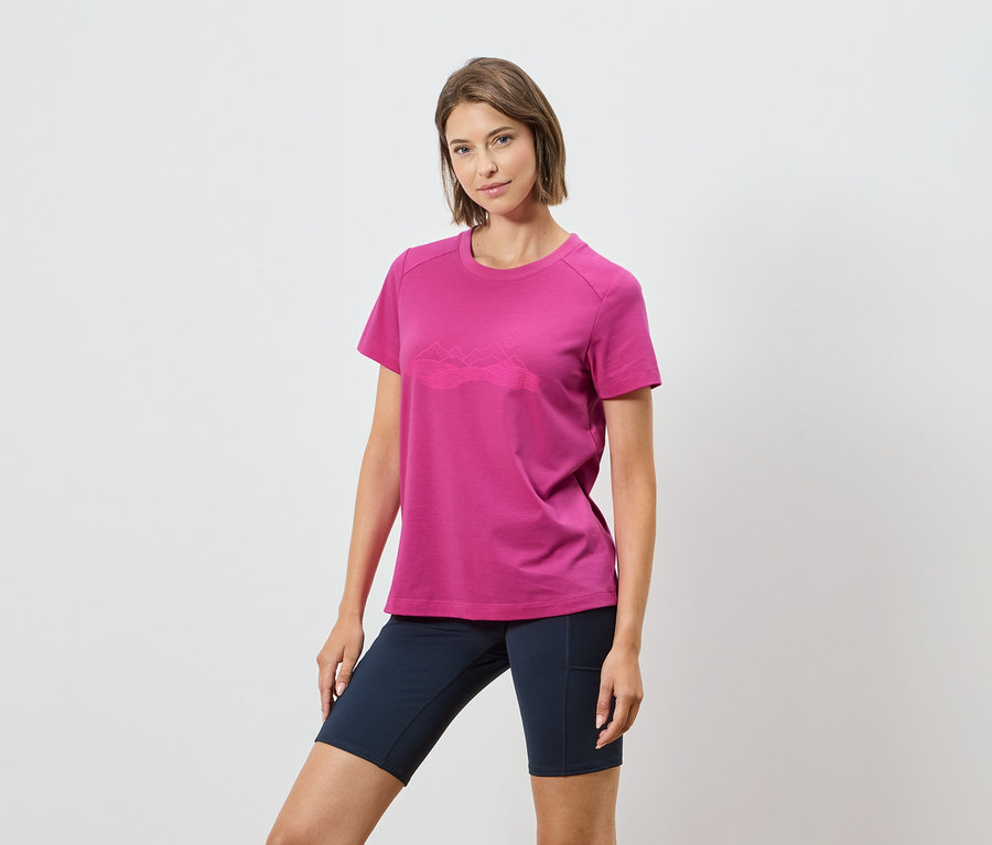 Egy nő rózsaszín pólót visel hegyvonulattal és sötétkék rövidnadrágot fehér háttér előtt.