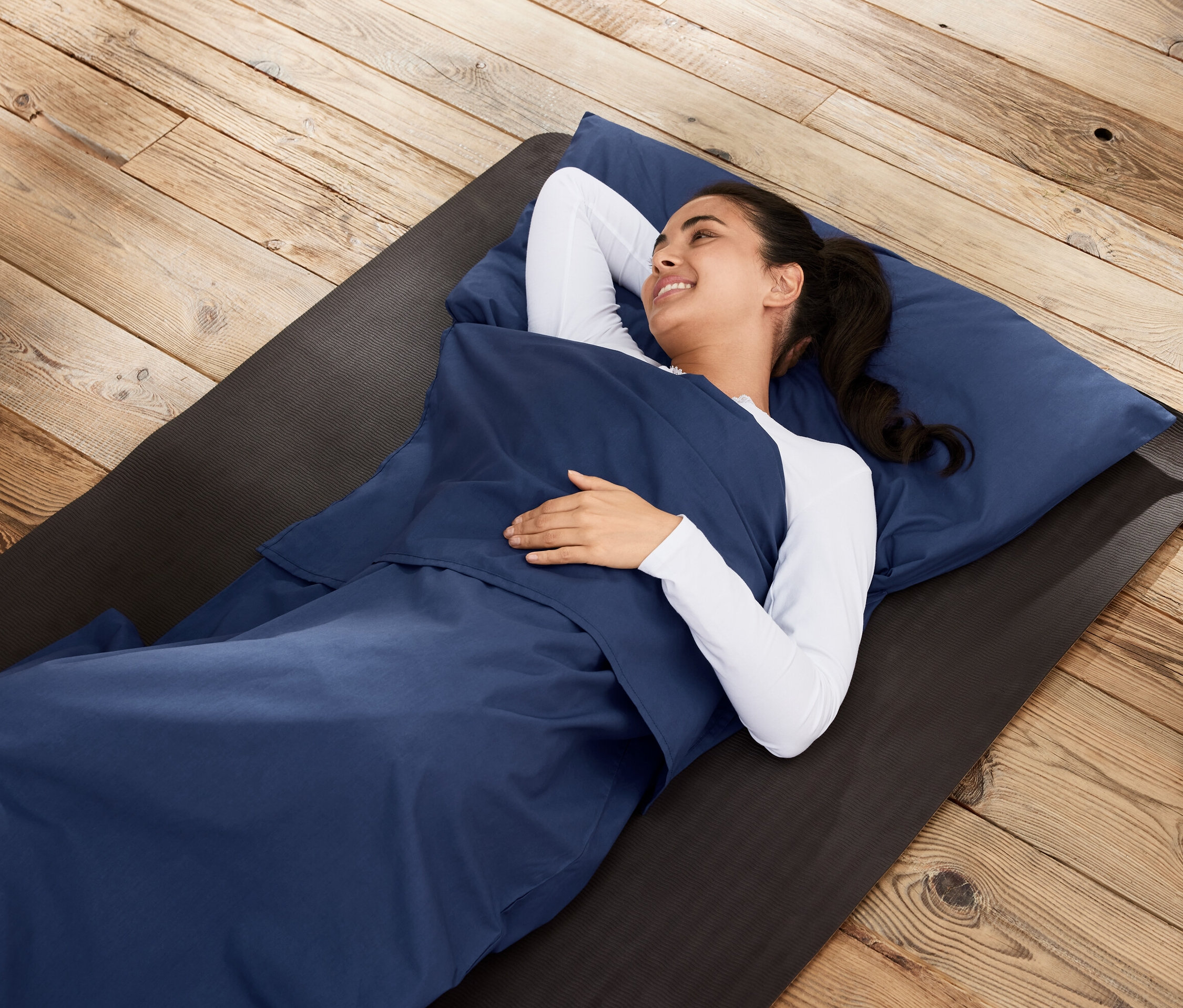 Nő fekszik hálózsákba való bélésben egy matracon, fapadlón.