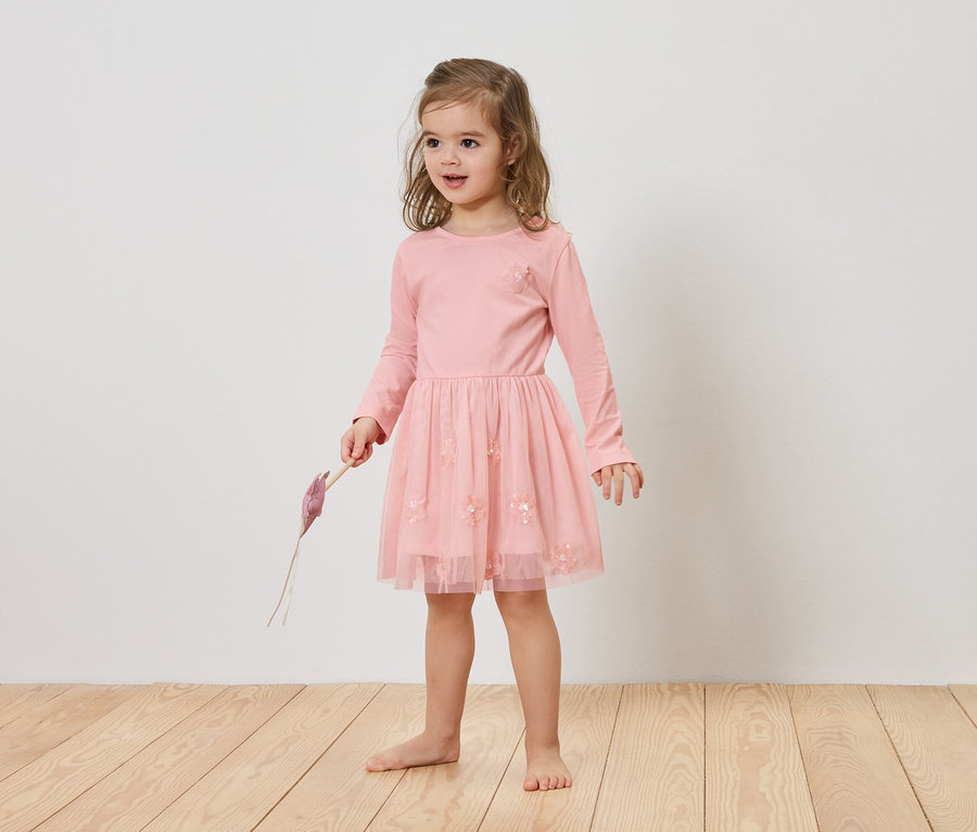 A szőke hajú lány rózsaszín ruhát visel és egy lila tárgyat tart a kezében.