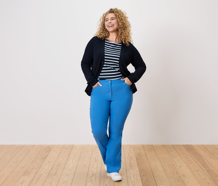 Göndör hajú nő visel sötétkék kardigánt, csíkos inget és kék nadrágot.