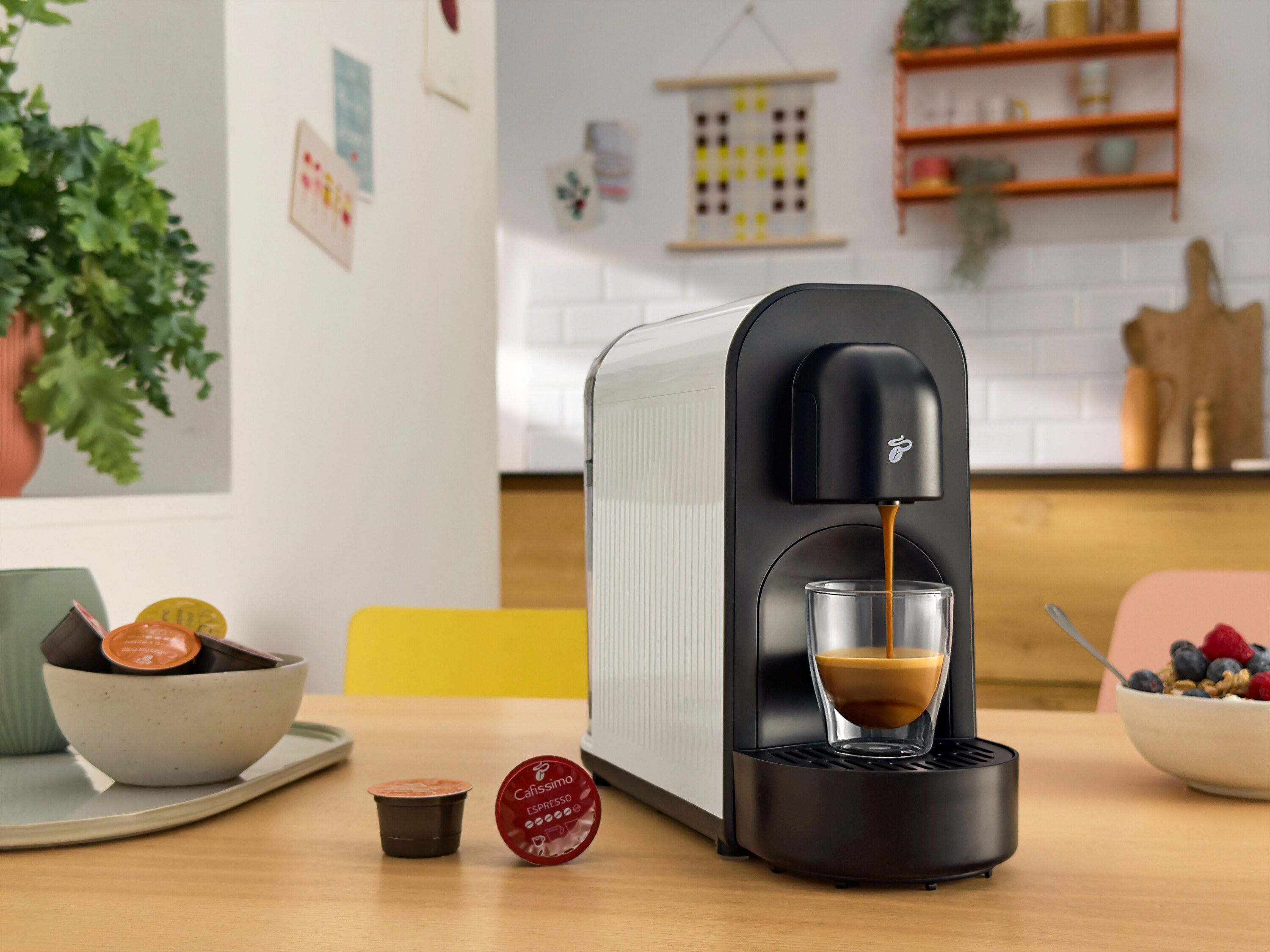 Kávé folyik a kávéfőzőből egy pohárba. Az asztalon kávékapszulák és müzlis tál található.