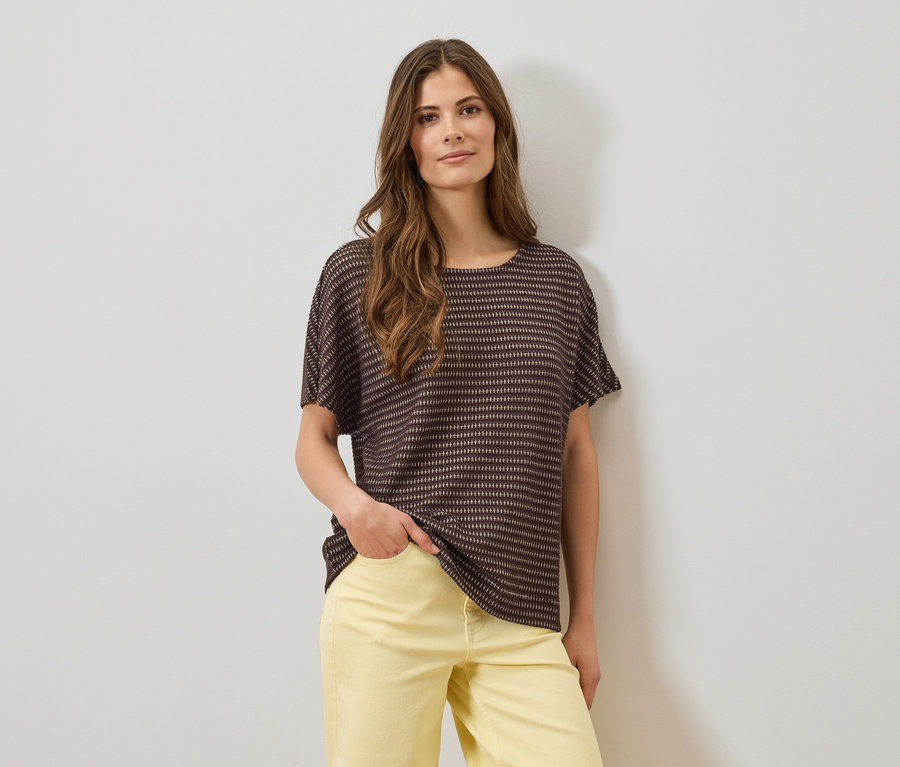 Nő barna, csíkos, rövid ujjú felsőben és sárga nadrágban, egyik kezével a zsebében, fehér fal előtt.