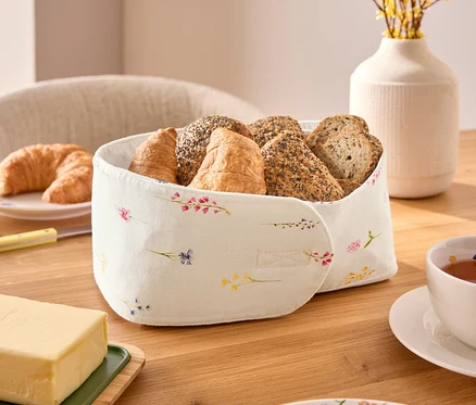 Egy fa asztalon egy kenyérkosár áll különféle pékárukkal, köztük croissant-okkal és zsemlékkel, egy csésze tea, egy darab vaj és egy virágmintás tányér mellett.