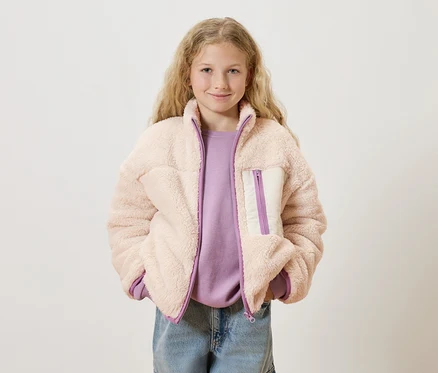 A szőke, göndör hajú lány rózsaszín teddy fleece kabátot visel lila pulóver és kék farmer felett.