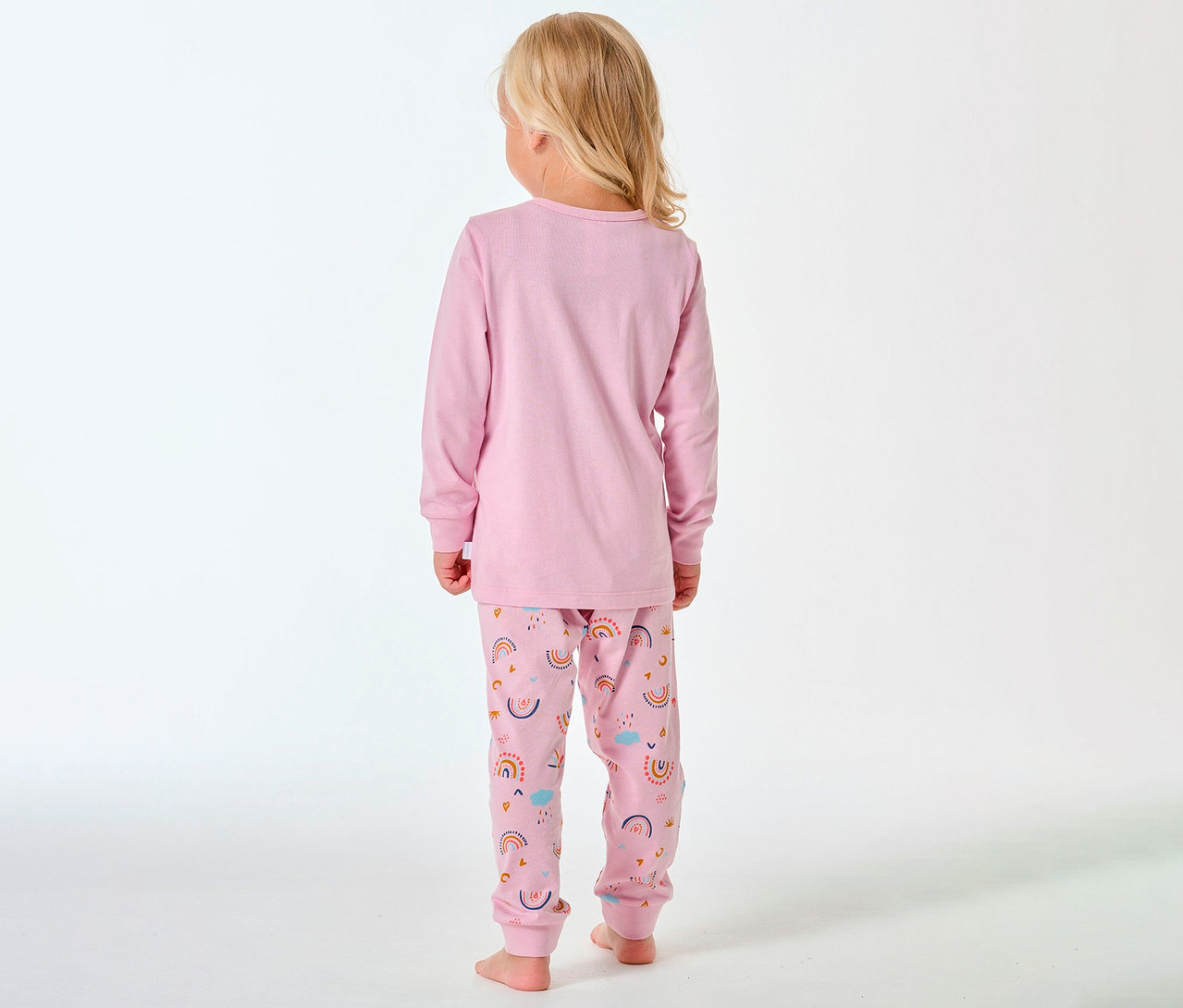 Hátulról látható, szőke hajú gyermek SCHIESSER hosszú gyerekpizsamát visel, szivárvány mintával.
