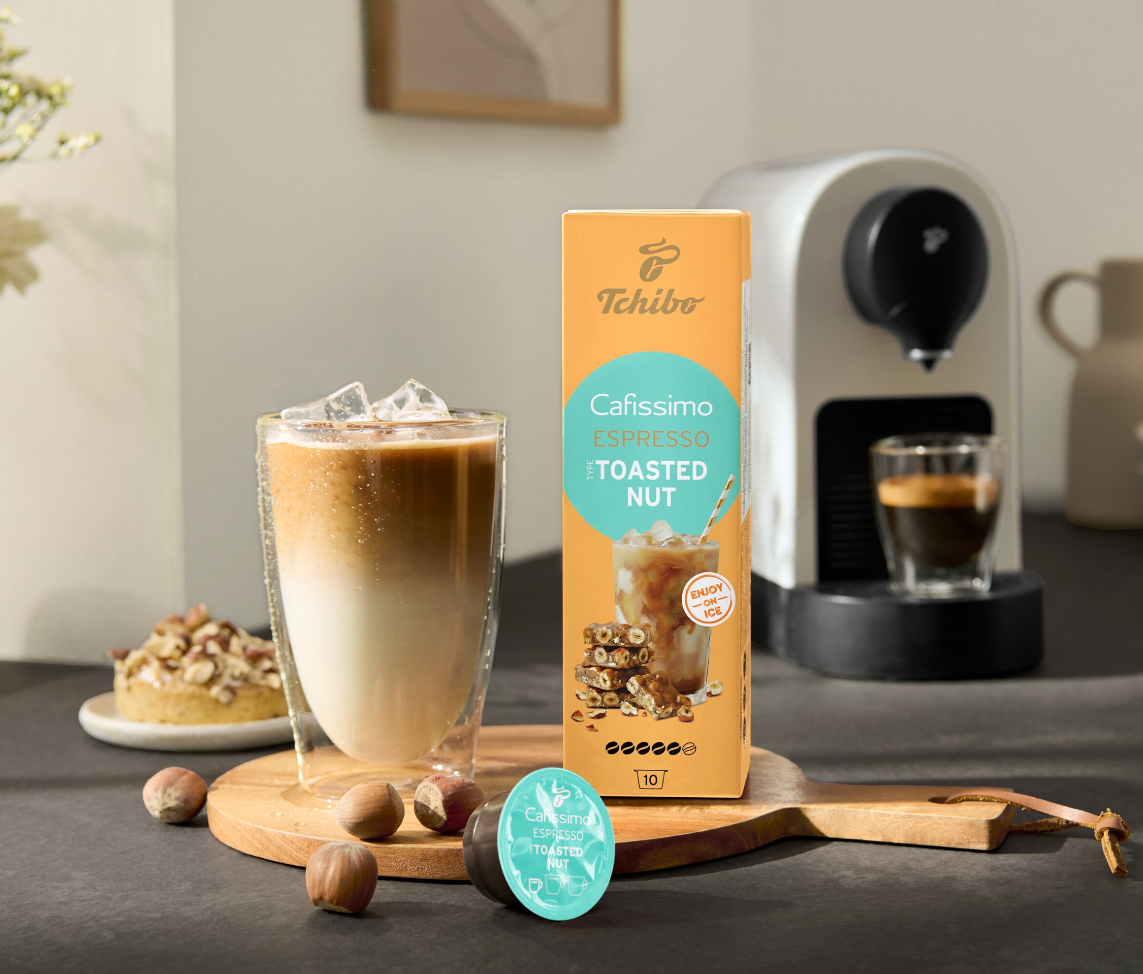 Egy pohár jeges kávé áll a Cafissimo Flavoured Espresso – Toasted Nut csomag és egy Cafissimo pure+, white kávéfőző mellett.