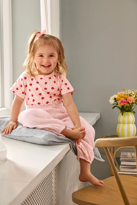 Egy mosolygó kislány ül egy párnán 2 Kinder-T-Shirts és Kinder-Musselin-Hose ruhában. Egy Váza van egy széken.