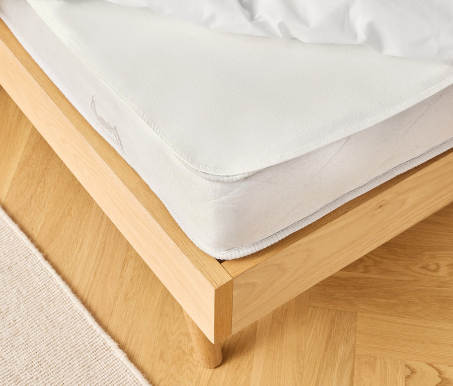 Egy ágy részletes képe fehér matraccal és ágyneművel világos fapadlón, mellette egy bézs szőnyeg.