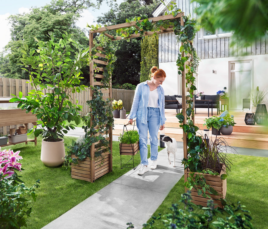 Egy nő egy kutyával sétál át egy növényekkel benőtt kerti boltíven. Egy virágládát visz.