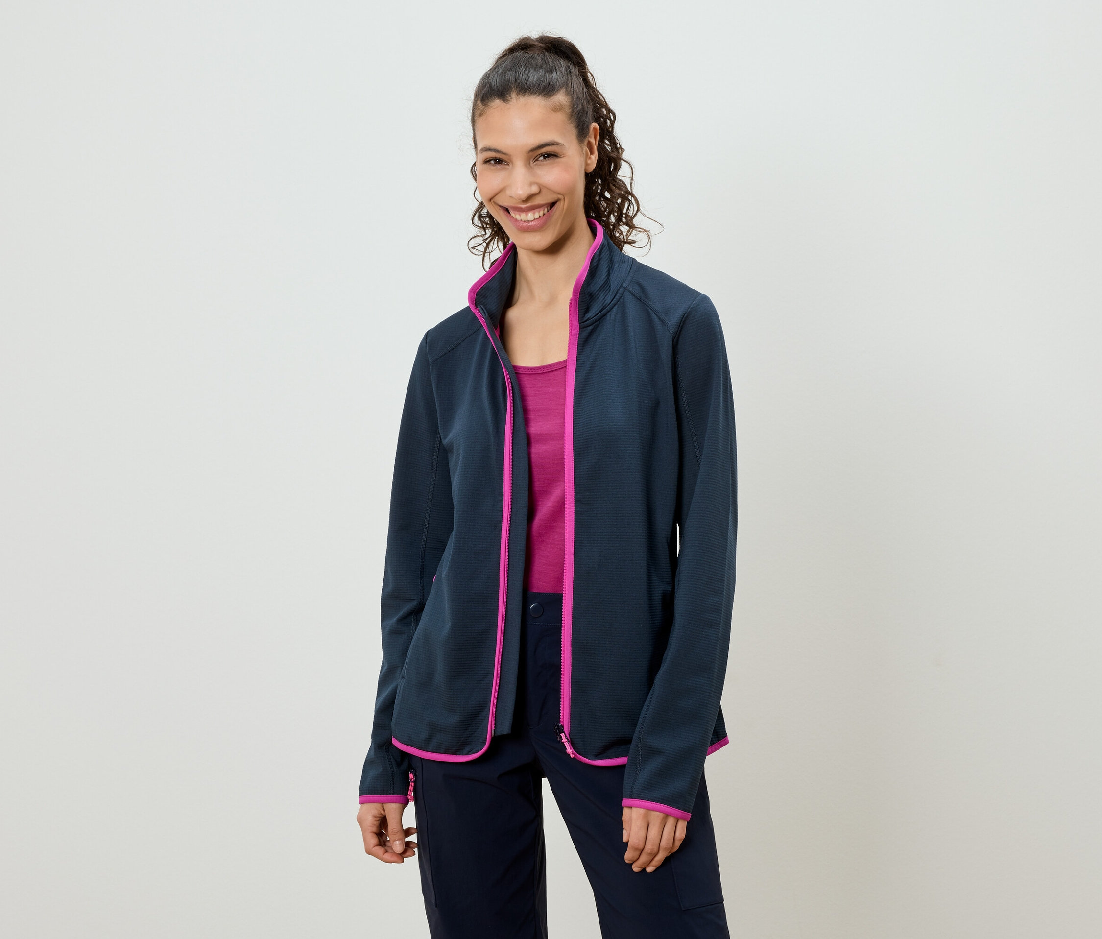 A lófarokba kötött hajú nő tengerészkék polár pulóvert visel rózsaszín díszítéssel, egy rózsaszín felsőt és tengerészkék nadrágot.