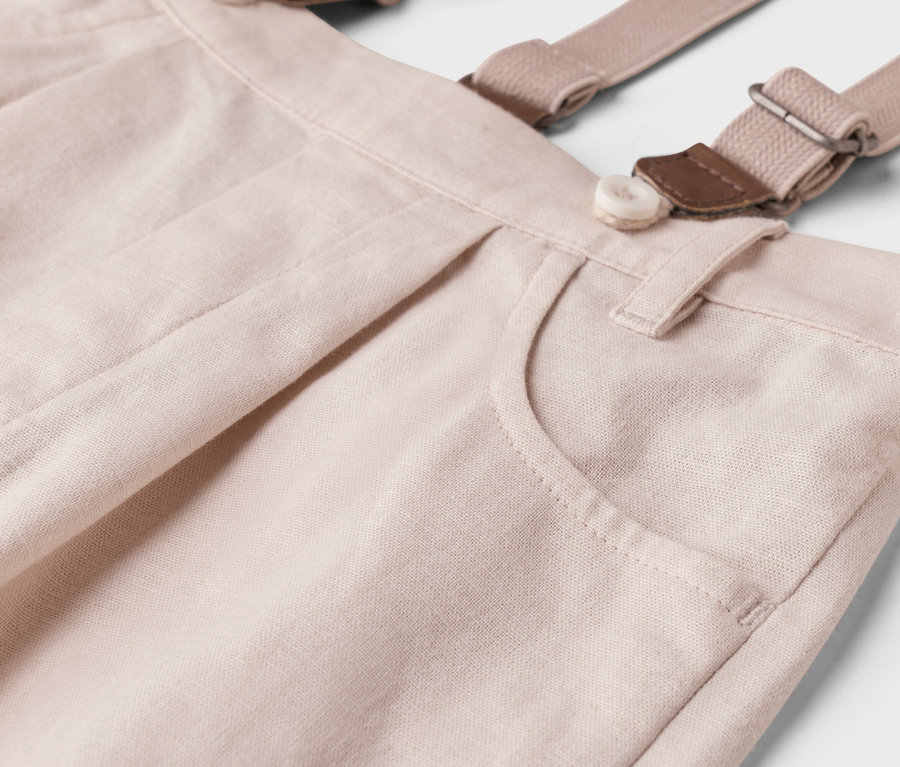 Közelkép egy világos bézs nadrágról, nadrágtartóval, gombbal és zsebbel.