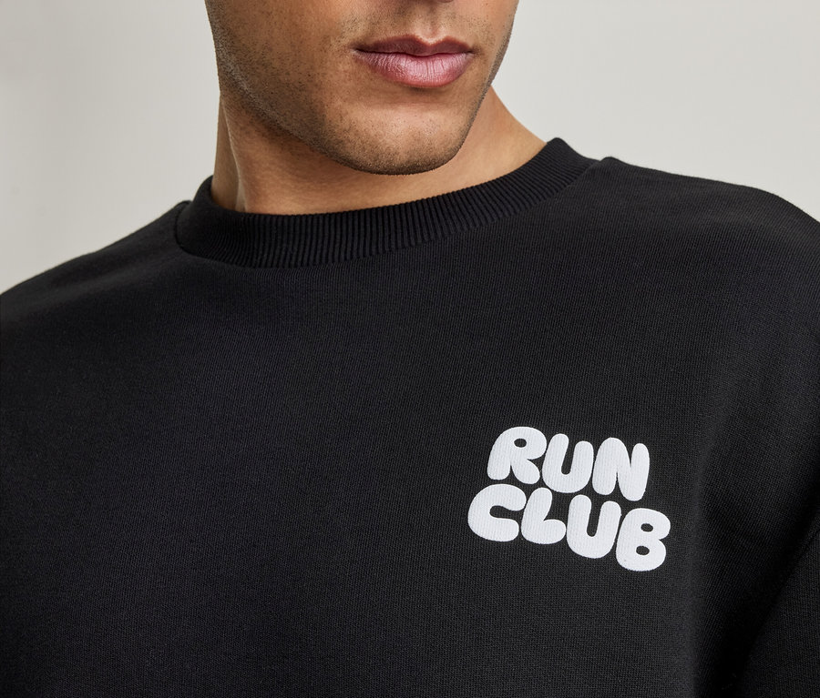 Férfi fekete szabadidőfelsőt visel, fehér 'RUN CLUB' felirattal, közelről.