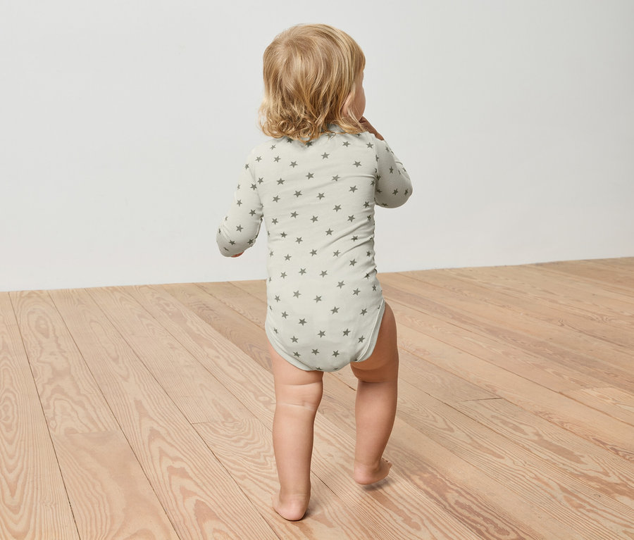 Egy szőke hajú kisgyermek csillagmintás világoszöld bodyt visel, és egy fapadlón sétál.