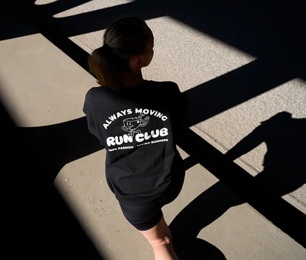 Nő hátulról fekete szabadidőfelsőben „Always Moving Run Club” felirattal és rövid, antracit »ActivePower« sportleggingsben, árnyékos úton.
