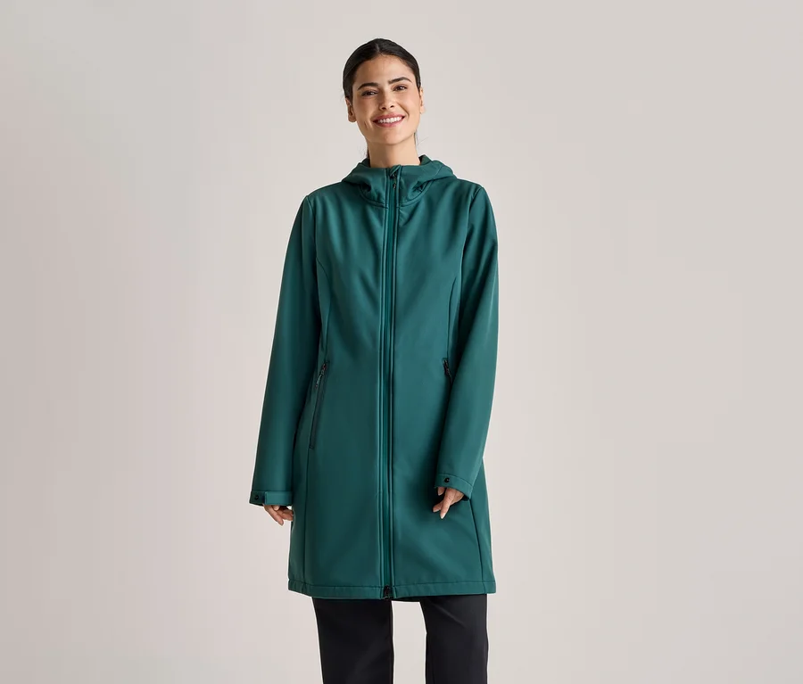 A lófarokba kötött hajú nő smaragdzöld, kapucnis, hosszú softshell kabátot és fekete nadrágot visel világosszürke háttér előtt.