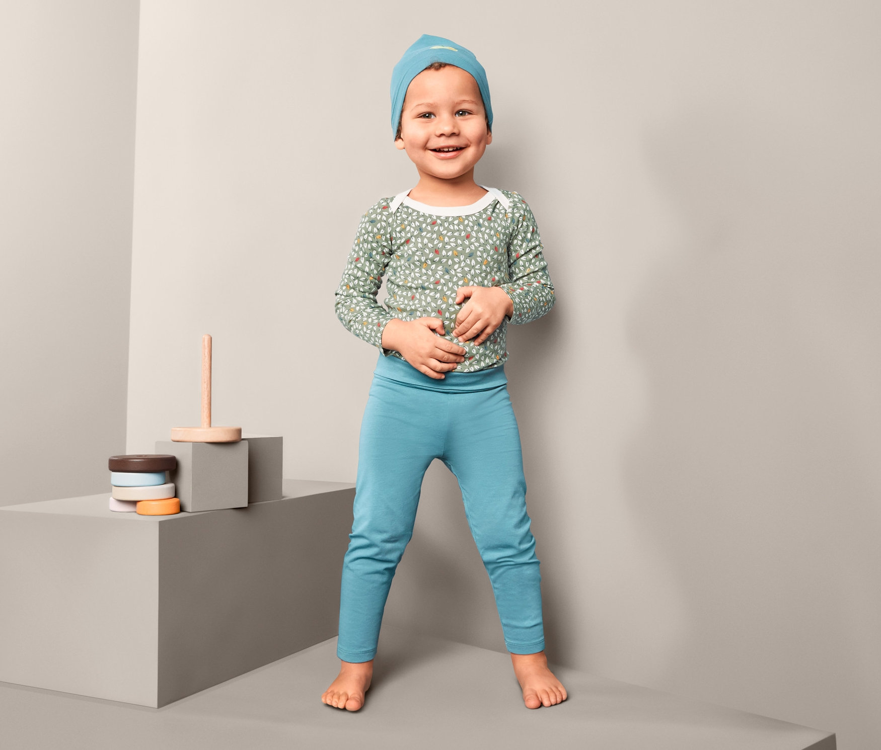 Egy mosolygó gyermek áll, és egy háromrészes kisgyerek együttest visel.