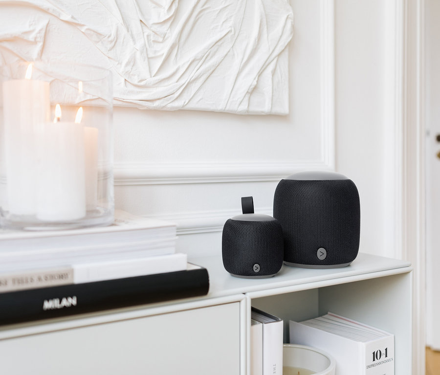 Két különböző méretű fekete Fabric Bluetooth® hangszóró áll egy fehér polcon gyertyák és könyvek mellett.
