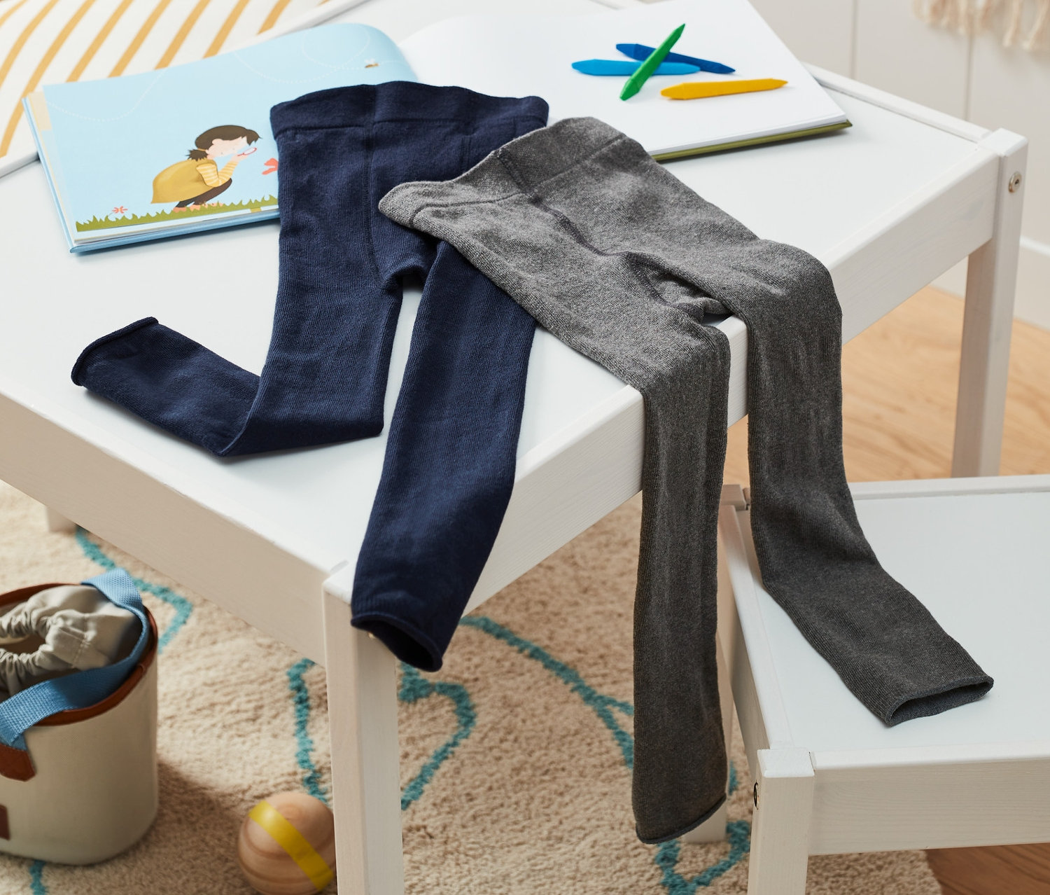 A fehér asztalon kék és szürke 2 gyerek kötött leggings, kék/szürke fekszik egy könyv és színes ceruzák mellett.