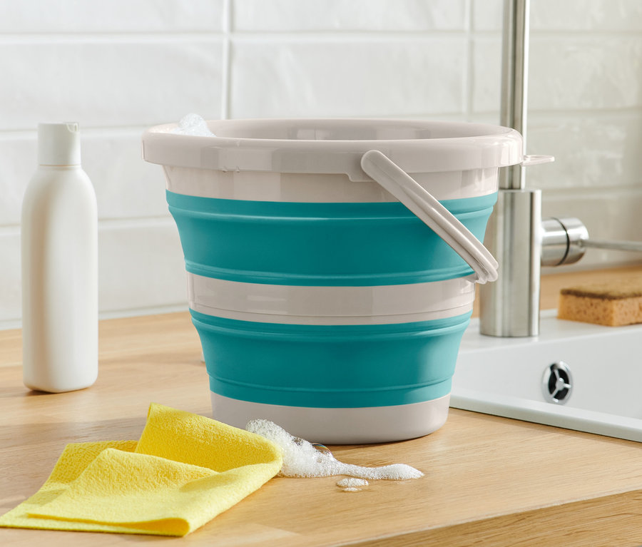 Egy összehajtható vödör áll egy fa deszkán egy sárga rongy és egy flakon mellett.