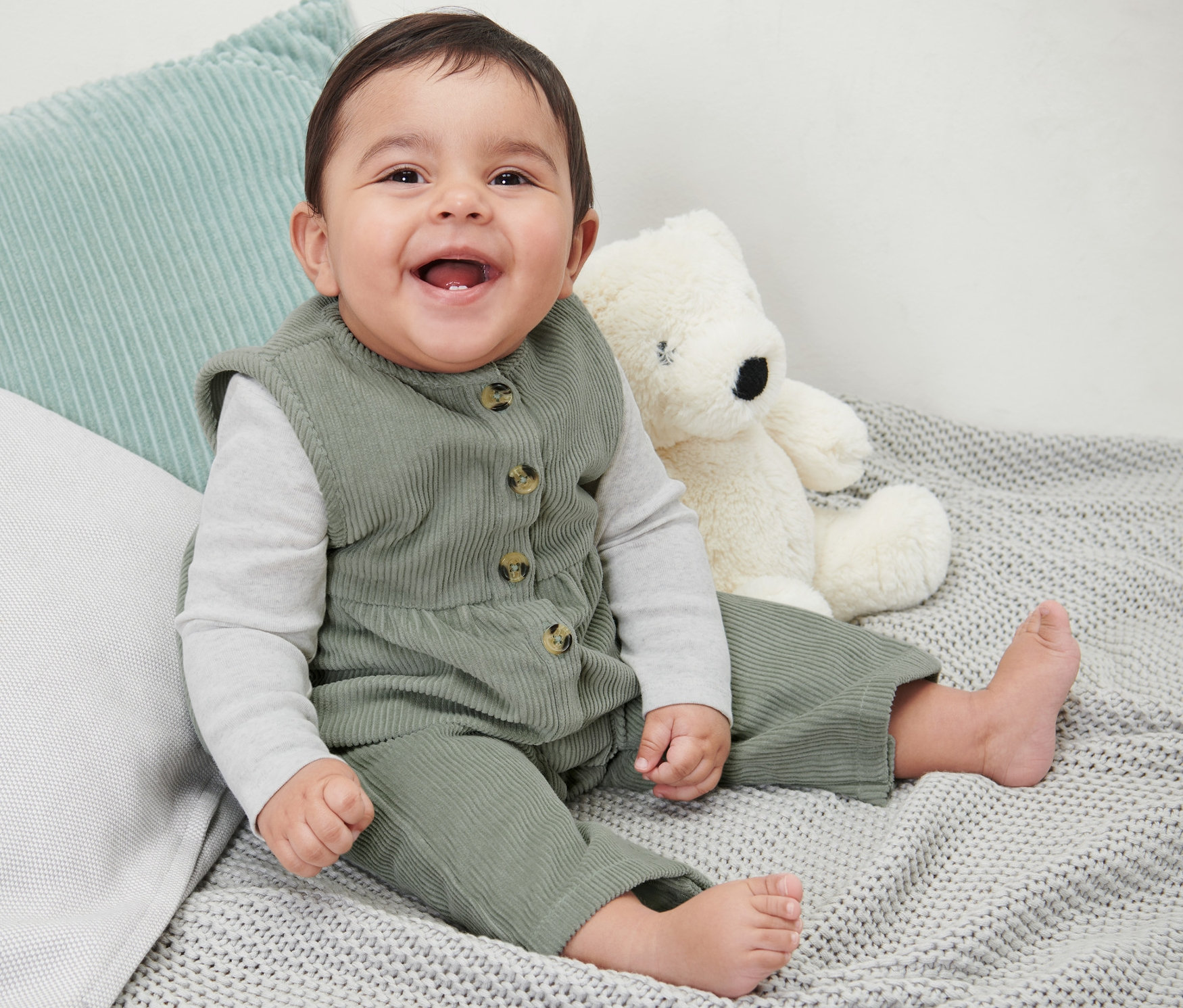 Egy mosolygó baba ül egy szürke kötött takarón egy plüssmackó mellett, és kordbársony kantáros nadrágot visel.