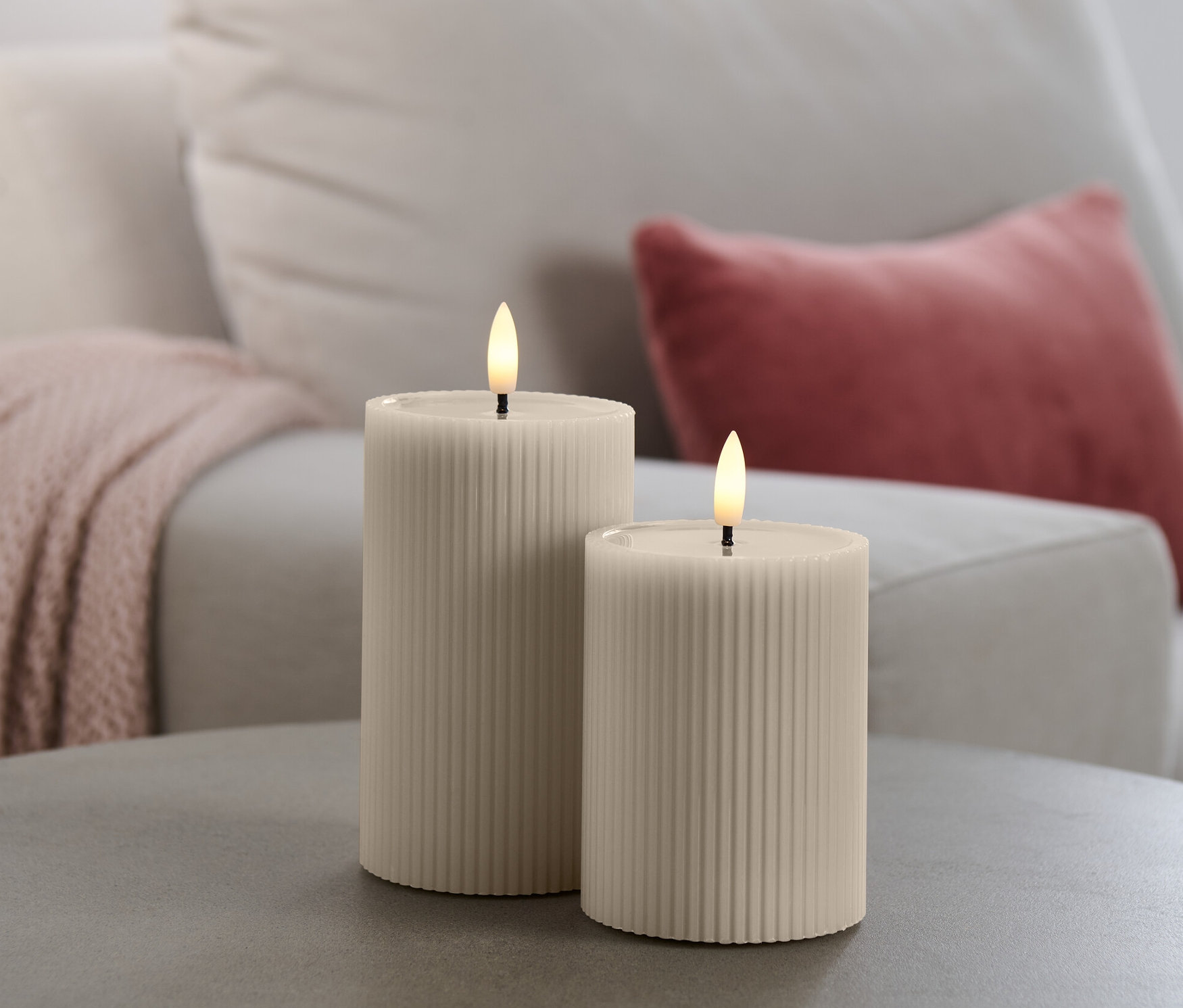 Két LED-es valódi viaszgyertya egy szürke asztalon, a háttérben egy kanapé.
