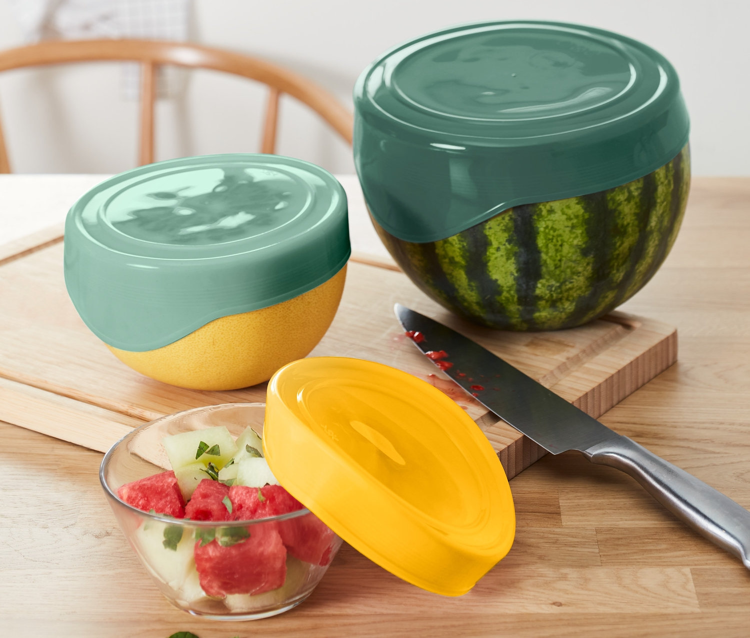 Két dinnye és egy üvegtál dinnyesaláta 3 szilikon frissentartó fedéllel van lefedve. Egy kés fekszik egy fa deszkán.