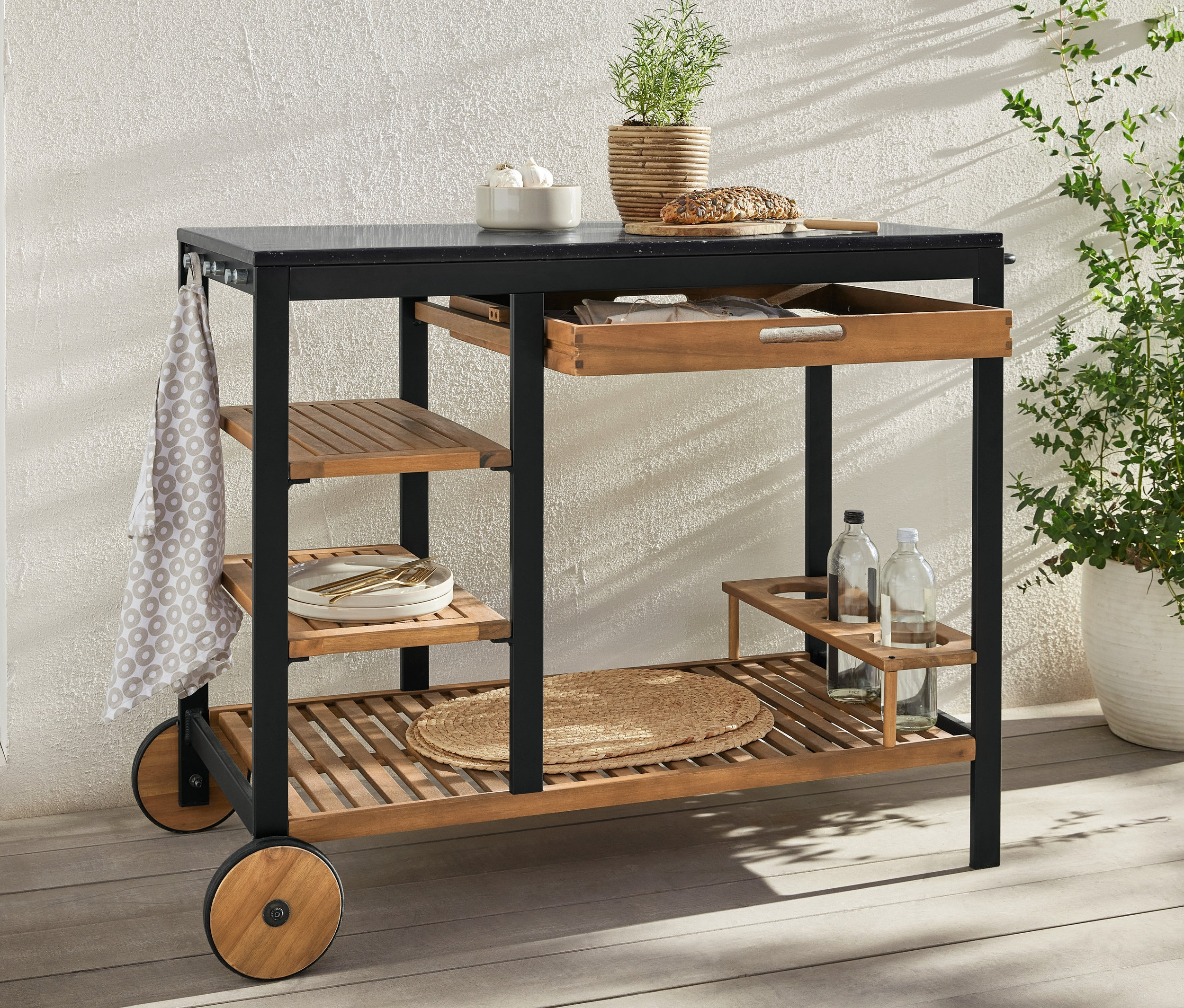 Egy „Elin” fiókos és cementhatású munkafelületes grillkocsi áll a szabadban. A munkafelületen kenyér, fokhagyma és egy növény található.