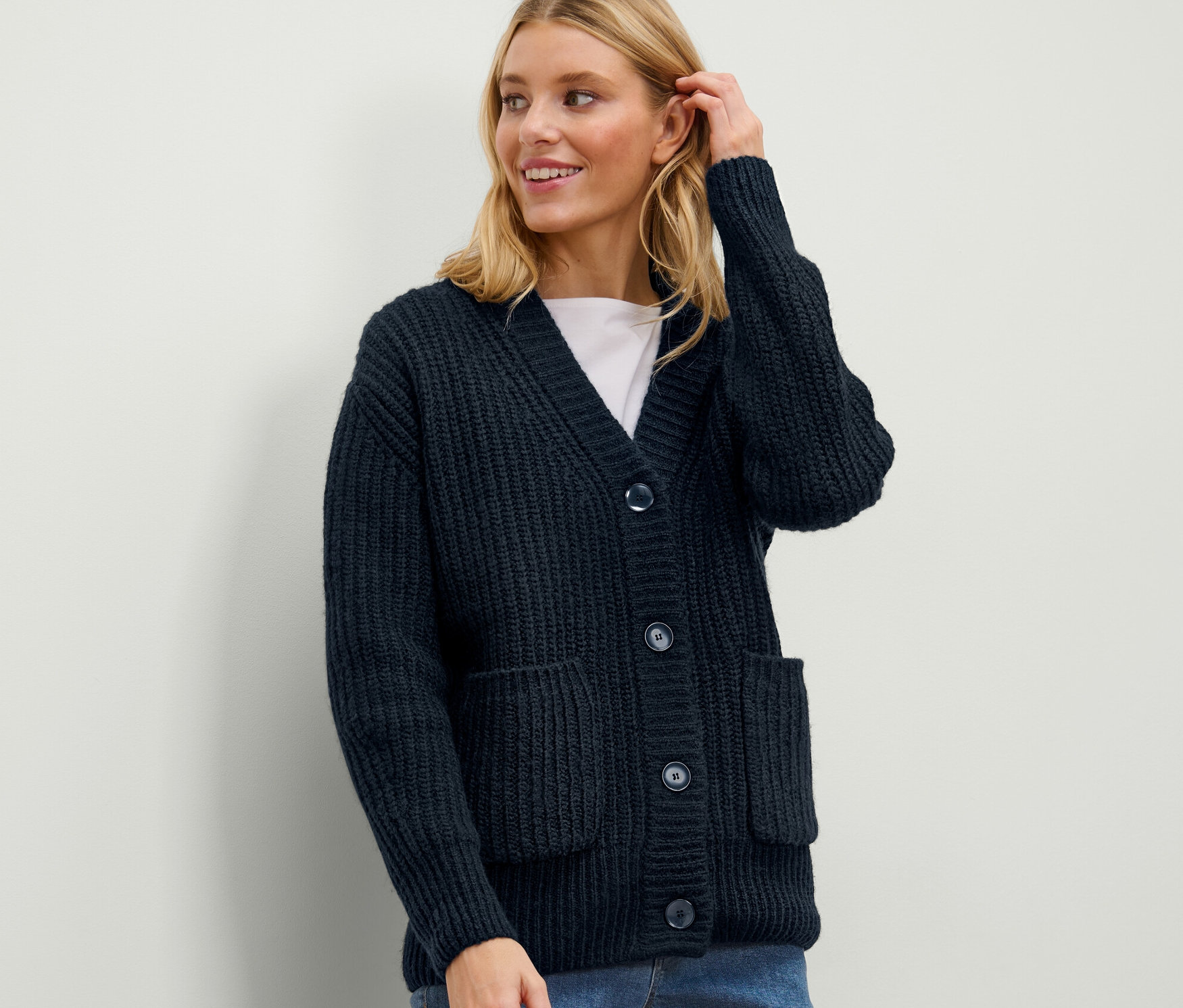 Egy szőke nő sötétkék, laza bordás kötésű kardigánt visel és megérinti a haját.