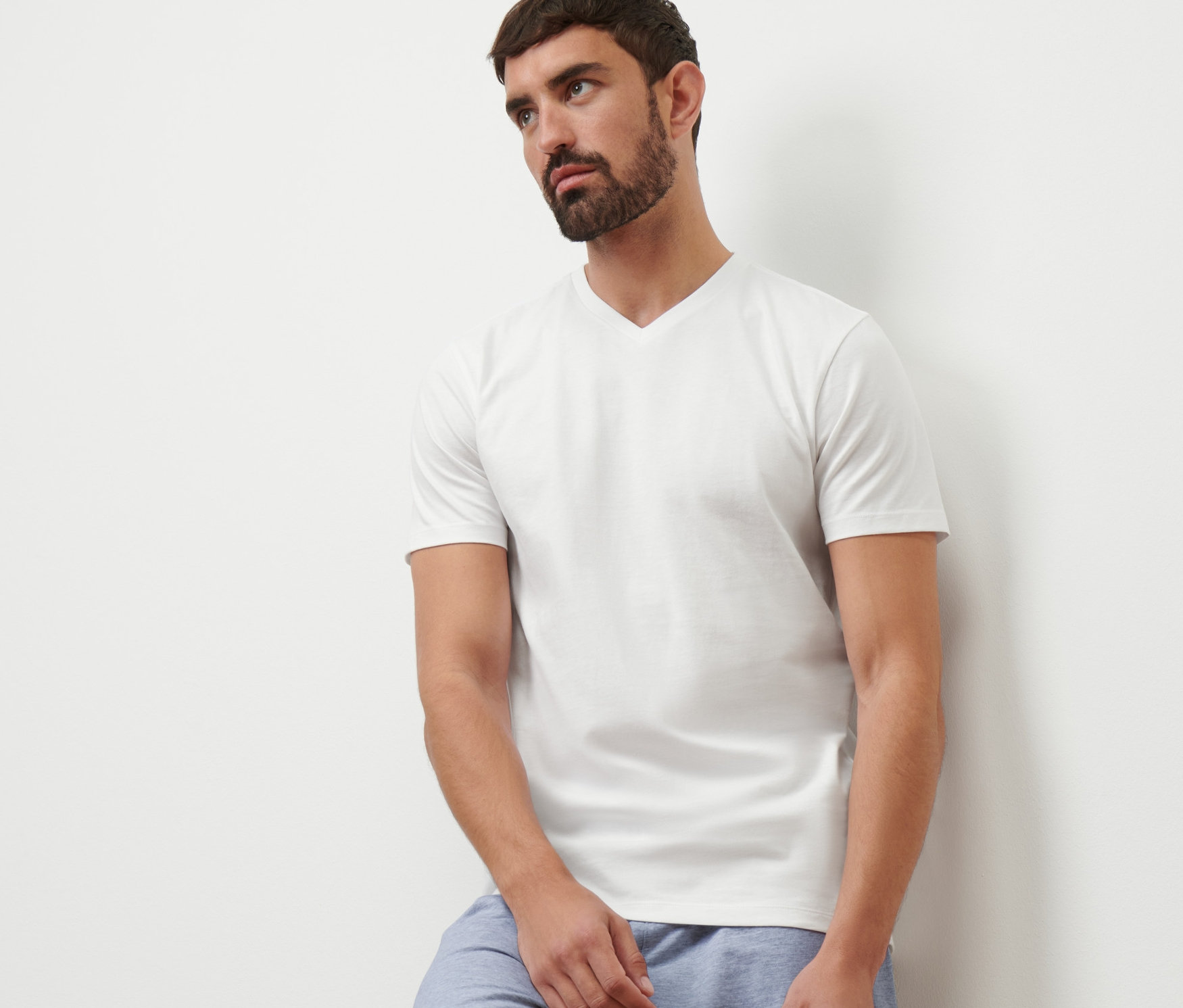 Egy falnak támaszkodó férfi fehér V-nyakú pólót és kék rövidnadrágot visel.