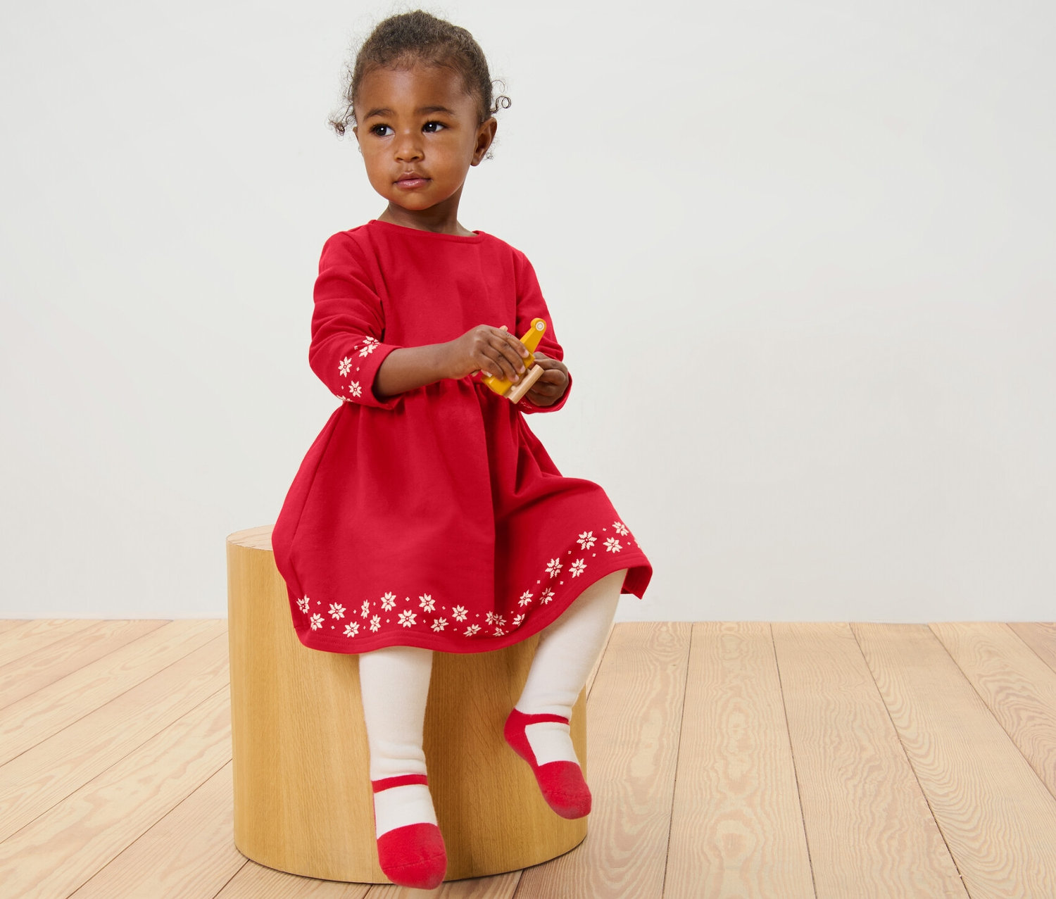 Egy kislány ül egy zsámolyon, és egy piros, mintás kislány lezser ruhát visel.