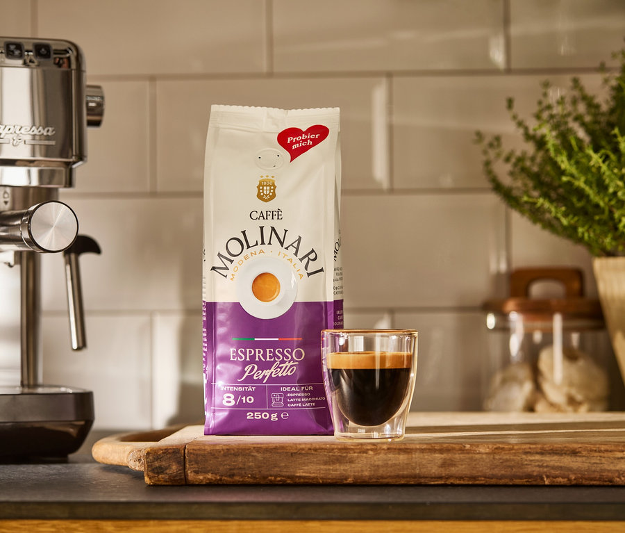 Egy ezüstszínű »Lapressa« karos kávéfőző, egy csomag Caffè Molinari és egy csésze eszpresszó egy fa deszkán.