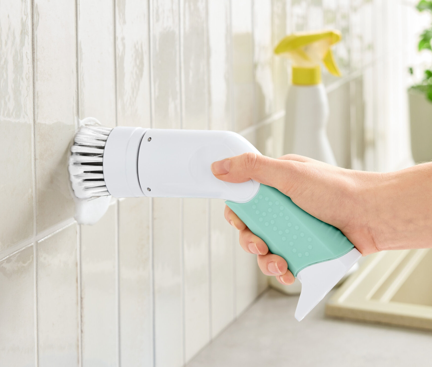 Egy kéz egy profi elektromos tisztítókefét tart, amivel fehér csempéket tisztít.