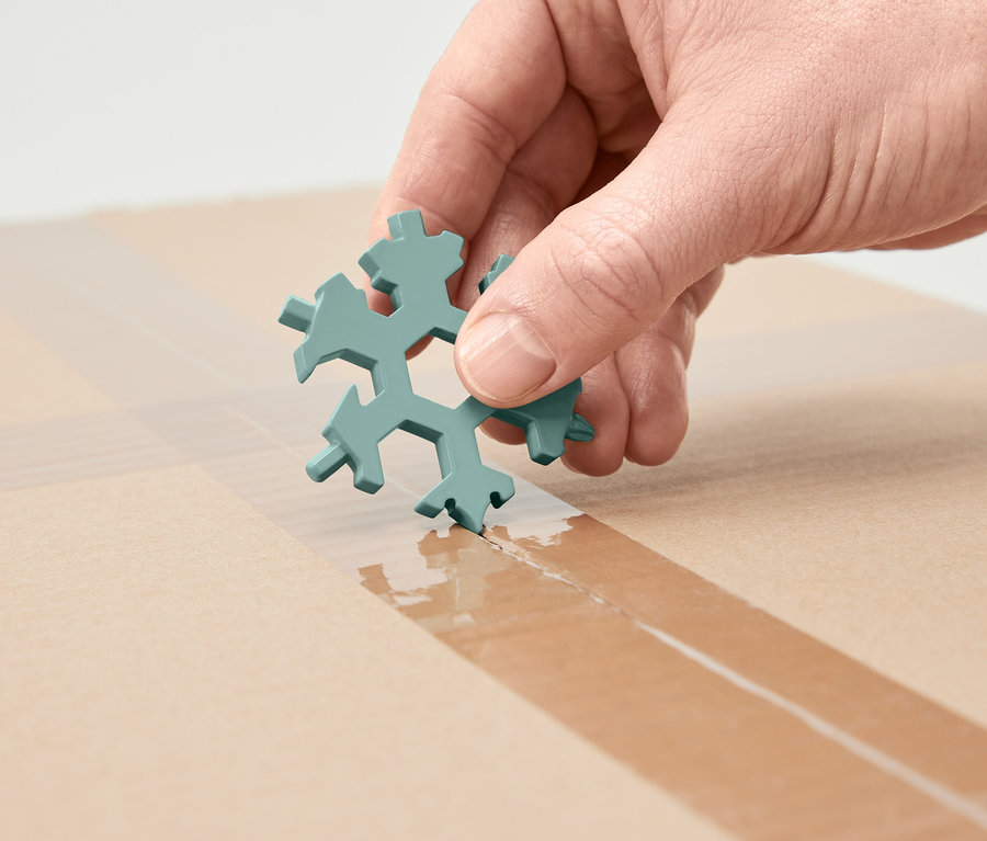 Egy kéz hópehely alakú multifunkciós kulcsot tart, és kartonon lévő ragasztószalagot vág vele.