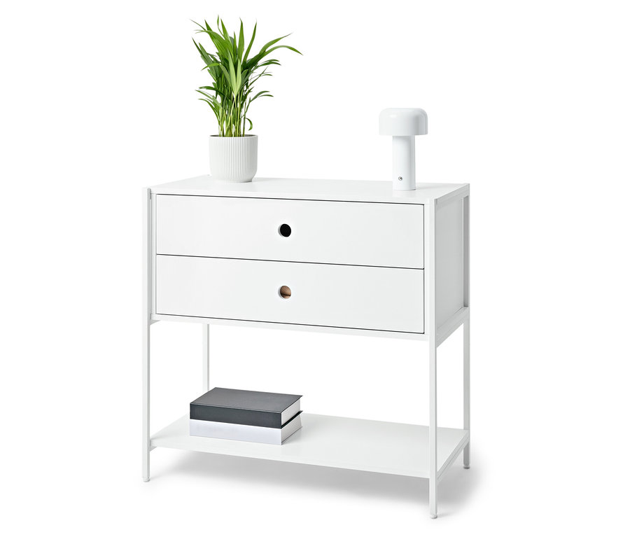 Fehér éjjeliszekrény két fiókkal, lámpával, cserepes növénnyel és könyvekkel.